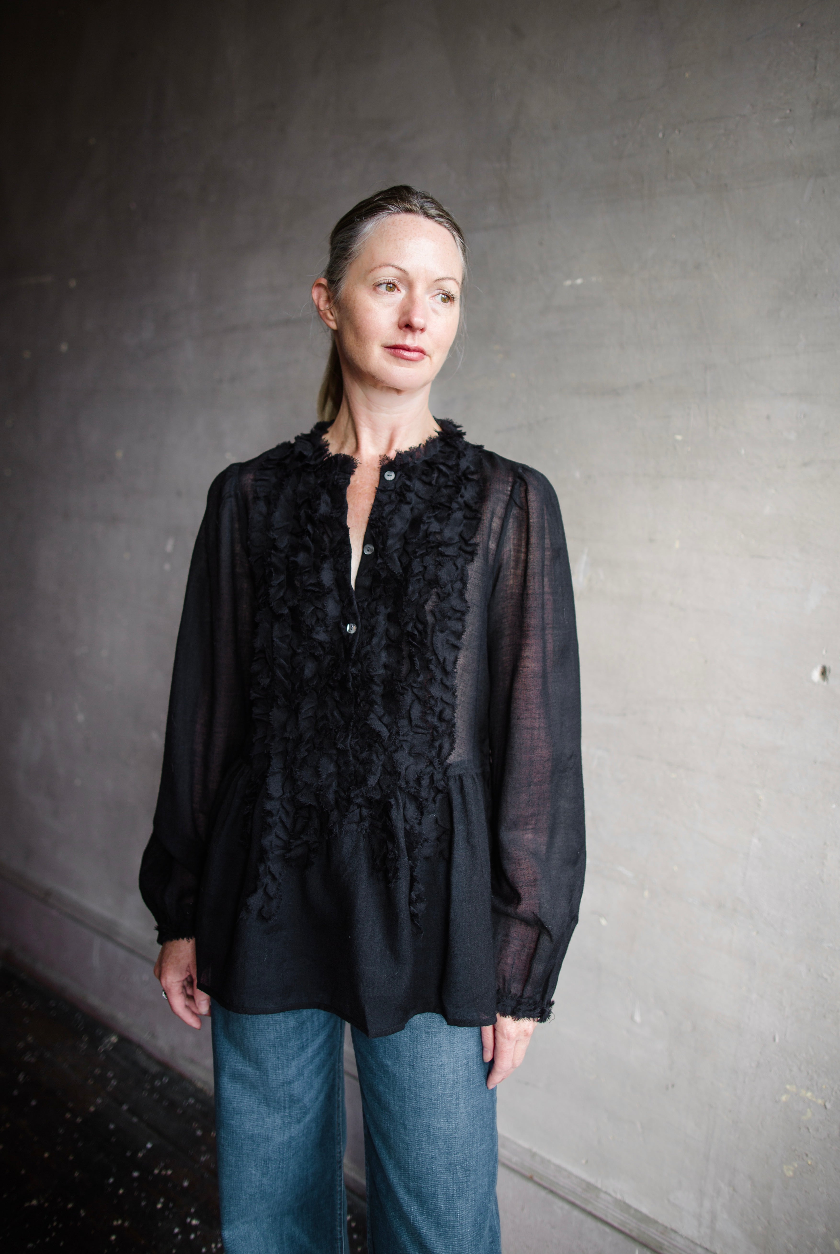 Image features woman wearing The Cosima Tunic by Ulla Johnson in Noir features a button down v neckline with rows of ruffles and iridescent sea shell buttons.