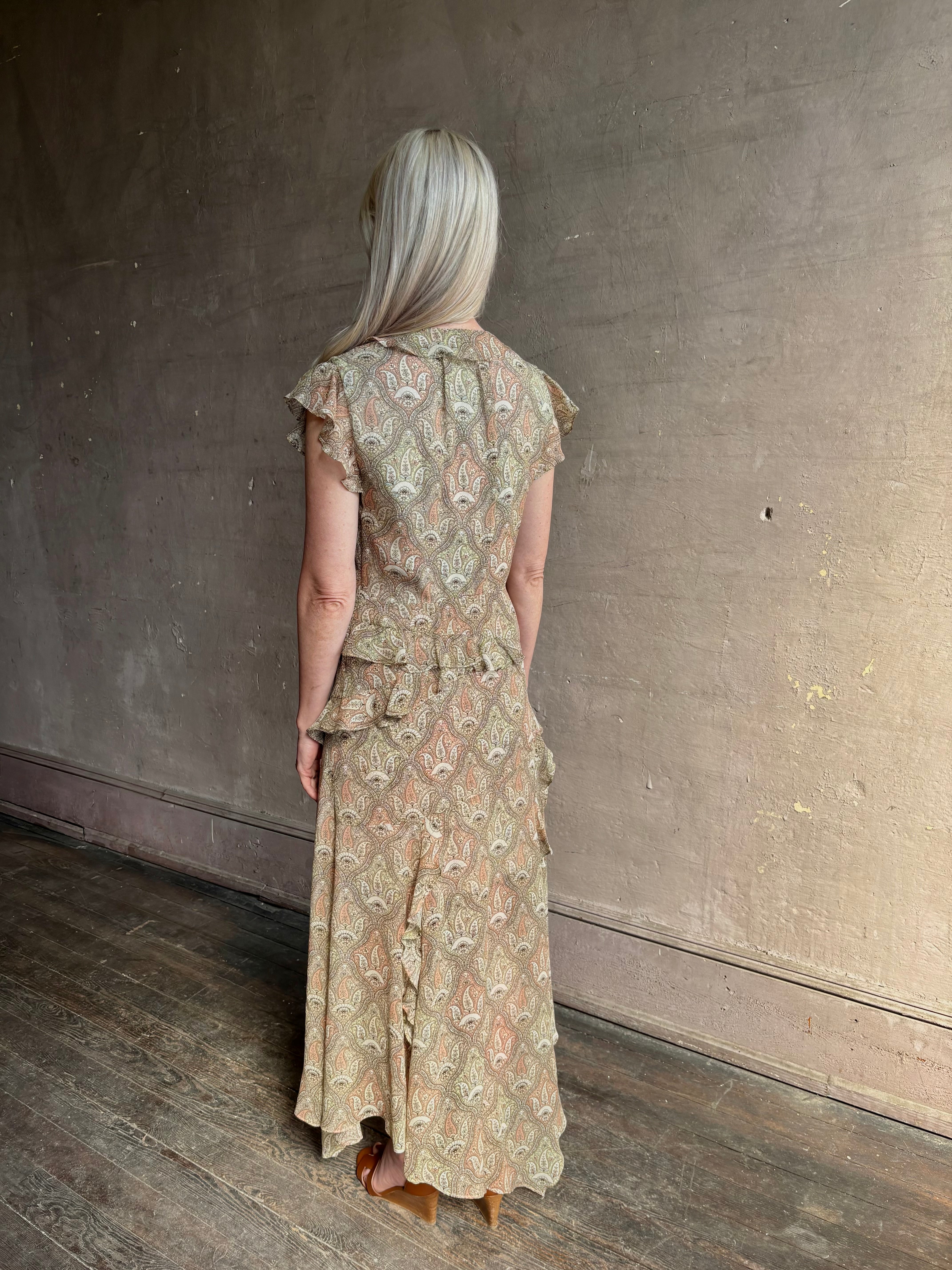 Mature woman wearing the Cap Sleeve Anenel maxi Dress from luxury designer Veronica Beard rendered in a soft khaki, green, and pink paisley print and crafted in silk with cap sleeves, v- neckline, and asymmetrical hem all finished with soft feminine ruffles in a timeless A-line silhouette