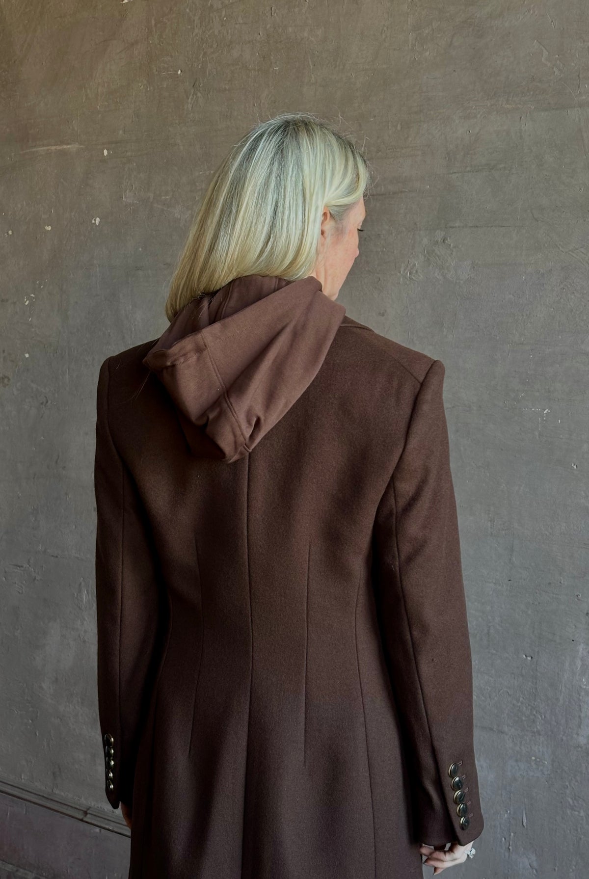 Mature woman wearing Veronica Beard Elspeth Coat with OVS Hoodie Dickey in Espresso brown wool-cashmere A-line coat with a removable hoodie layer. Playful, sculptural cold-weather luxury.