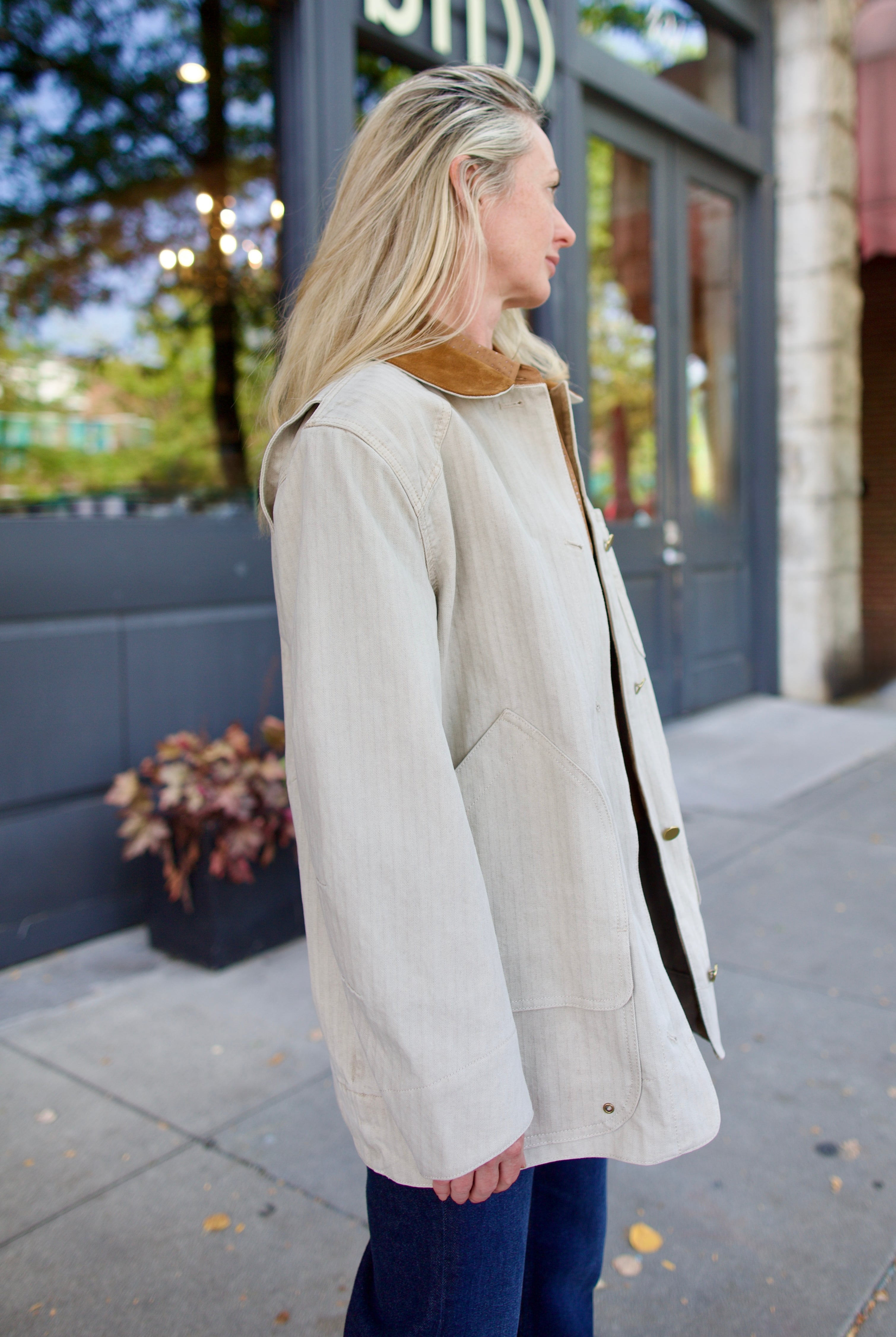 Mature model wearing a casual light weight cotton neutral barn jacket with a suede collar and denim.