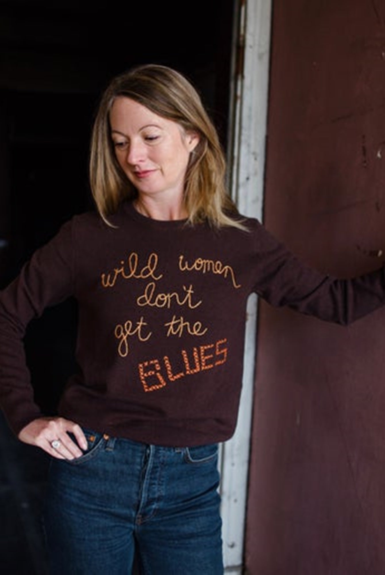 Image featuring a woman wearing the brown cashmere sweater by Lingua Franca with a crew neckline, long sleeves, and "Wild women don't get the blues" embroidered on front.