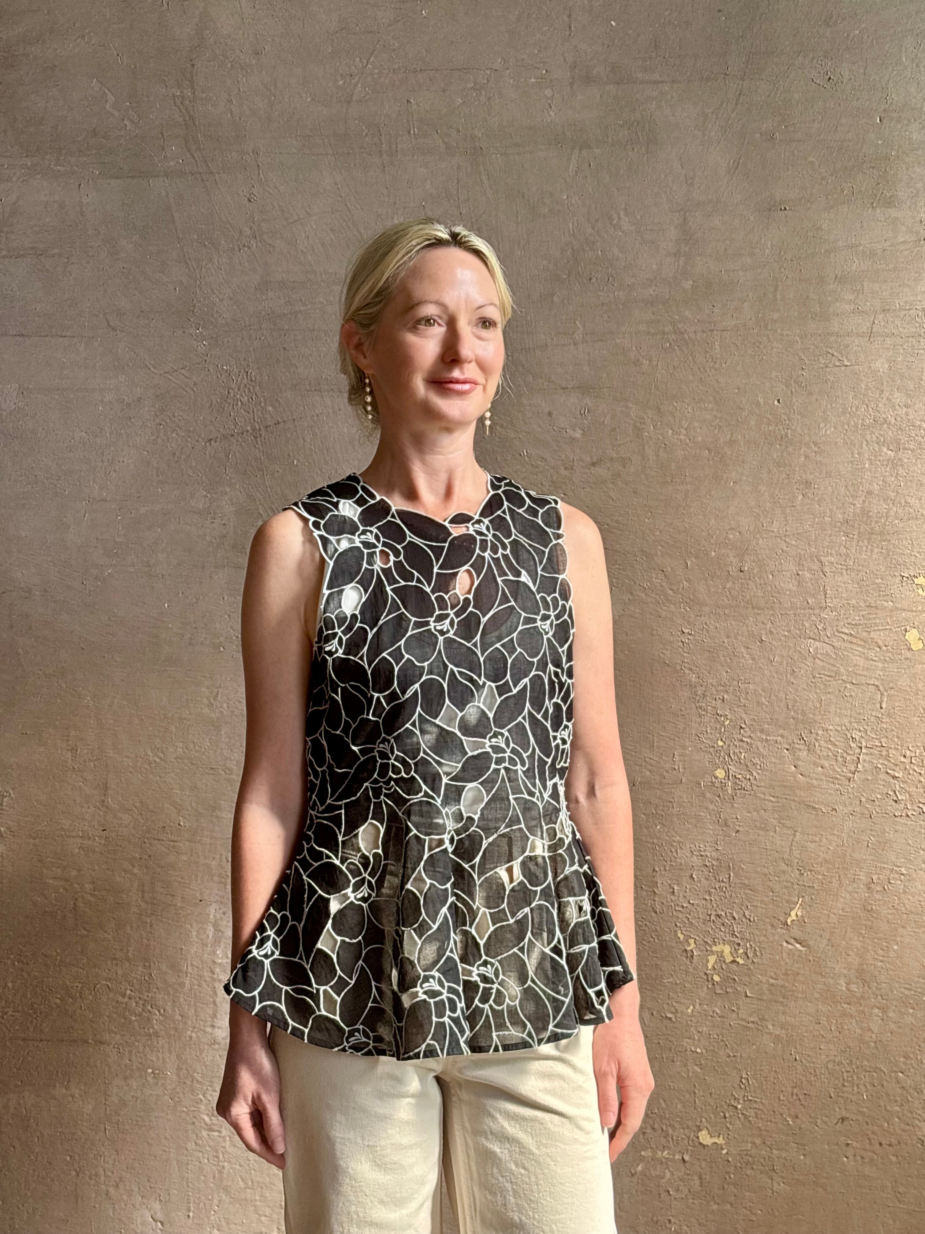 Mature woman wearing the Ulla Johnson Ali Sleeveless black cotton peplum top with contrast white embroidery for ultimate feminine sophistication