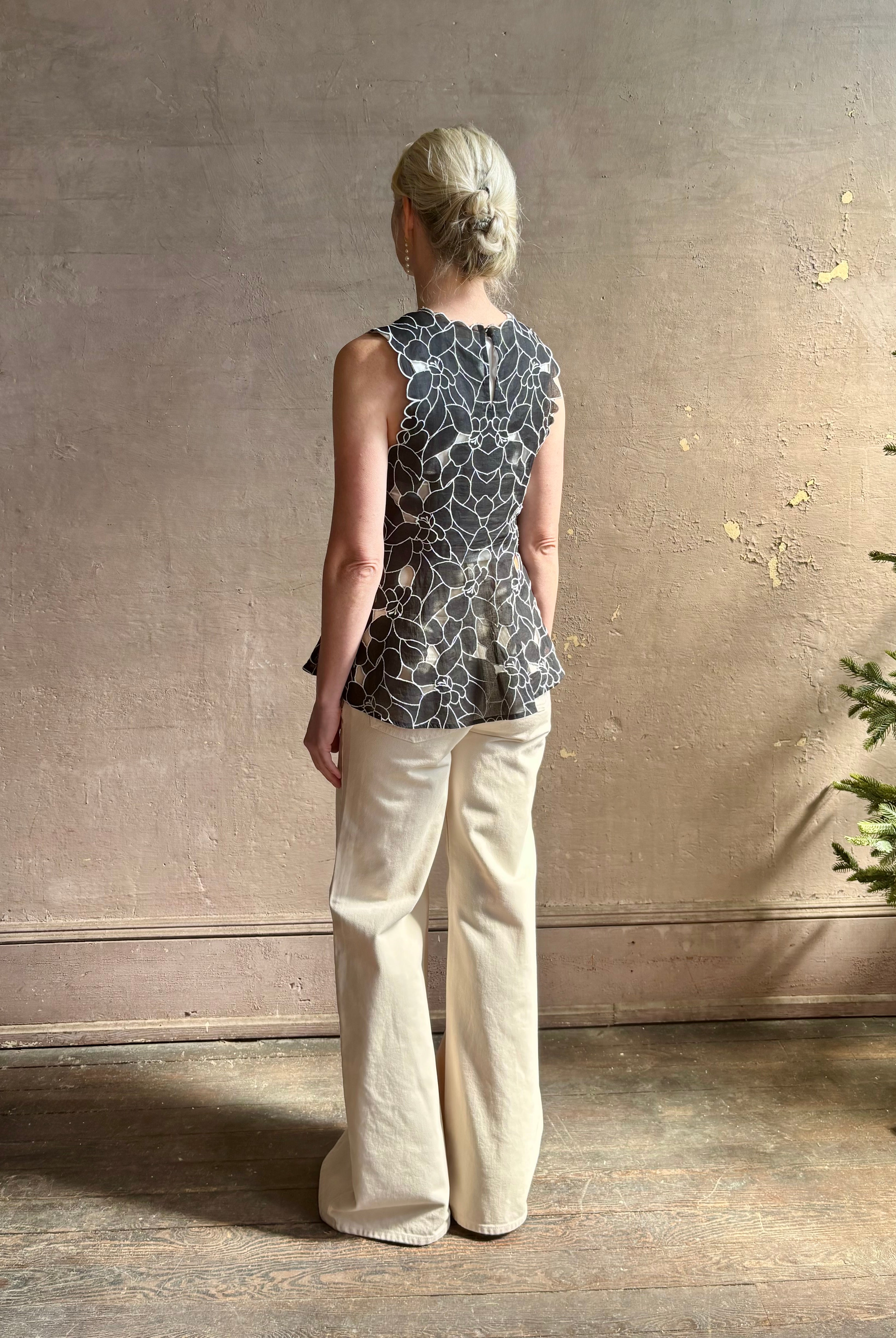 Mature woman wearing the Ulla Johnson Ali Sleeveless black cotton peplum top with contrast white embroidery for ultimate feminine sophistication