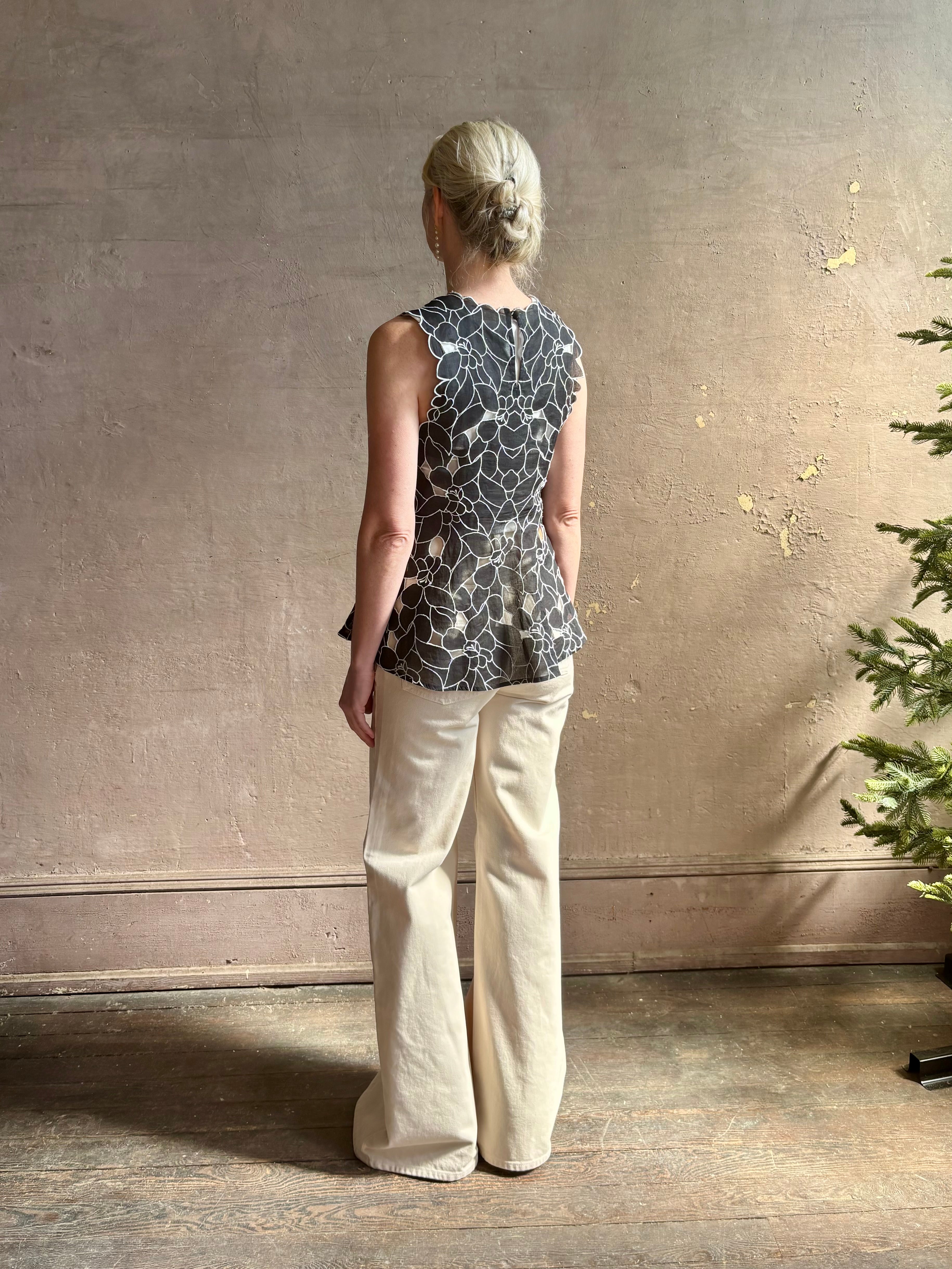 Mature woman wearing the Ulla Johnson Ali Sleeveless black cotton peplum top with contrast white embroidery for ultimate feminine sophistication