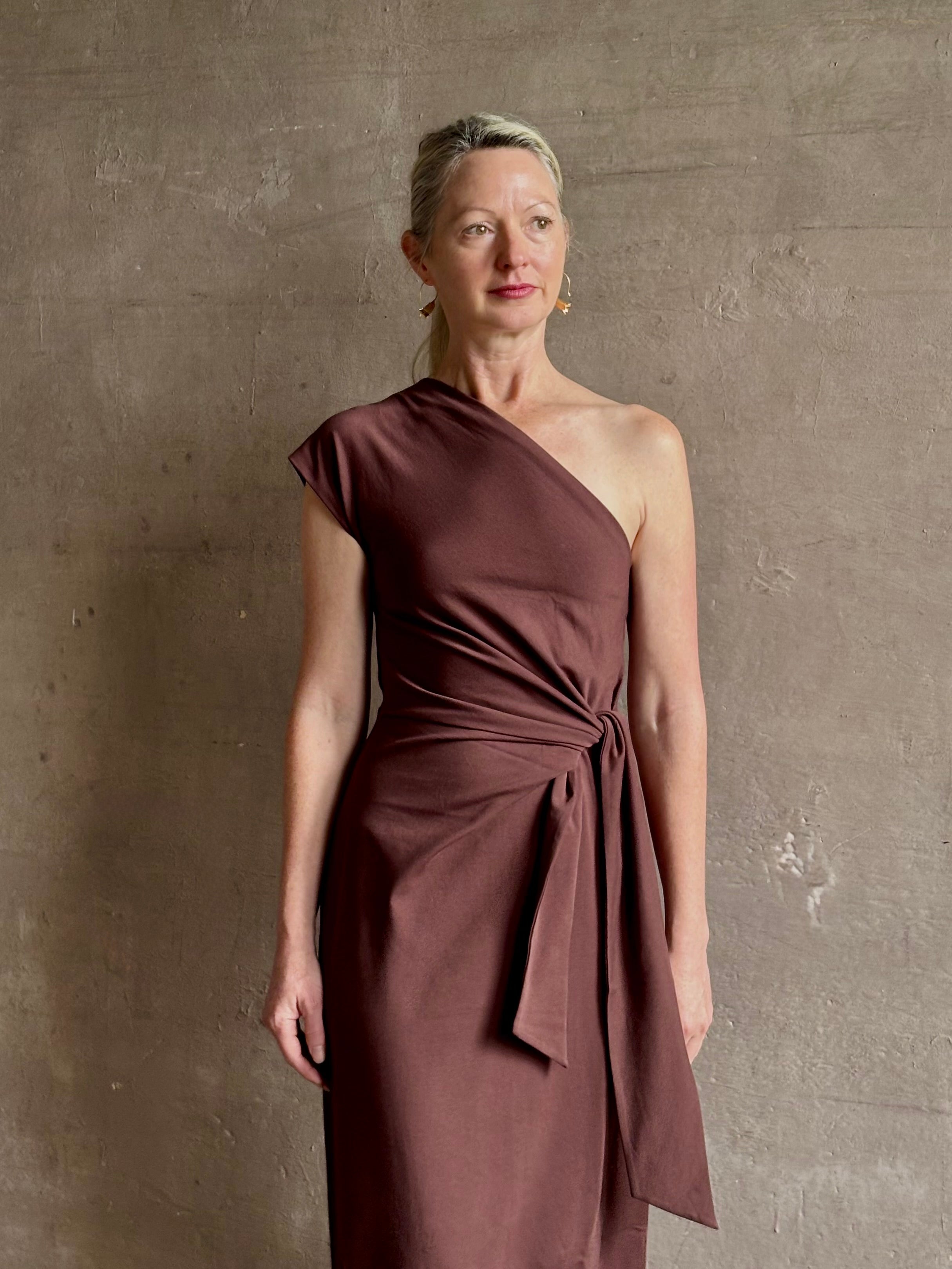 Mature woman wearing Apiece Apart brown comfy jersey cotton maxi dress one shoulder twist tie waist stretch
