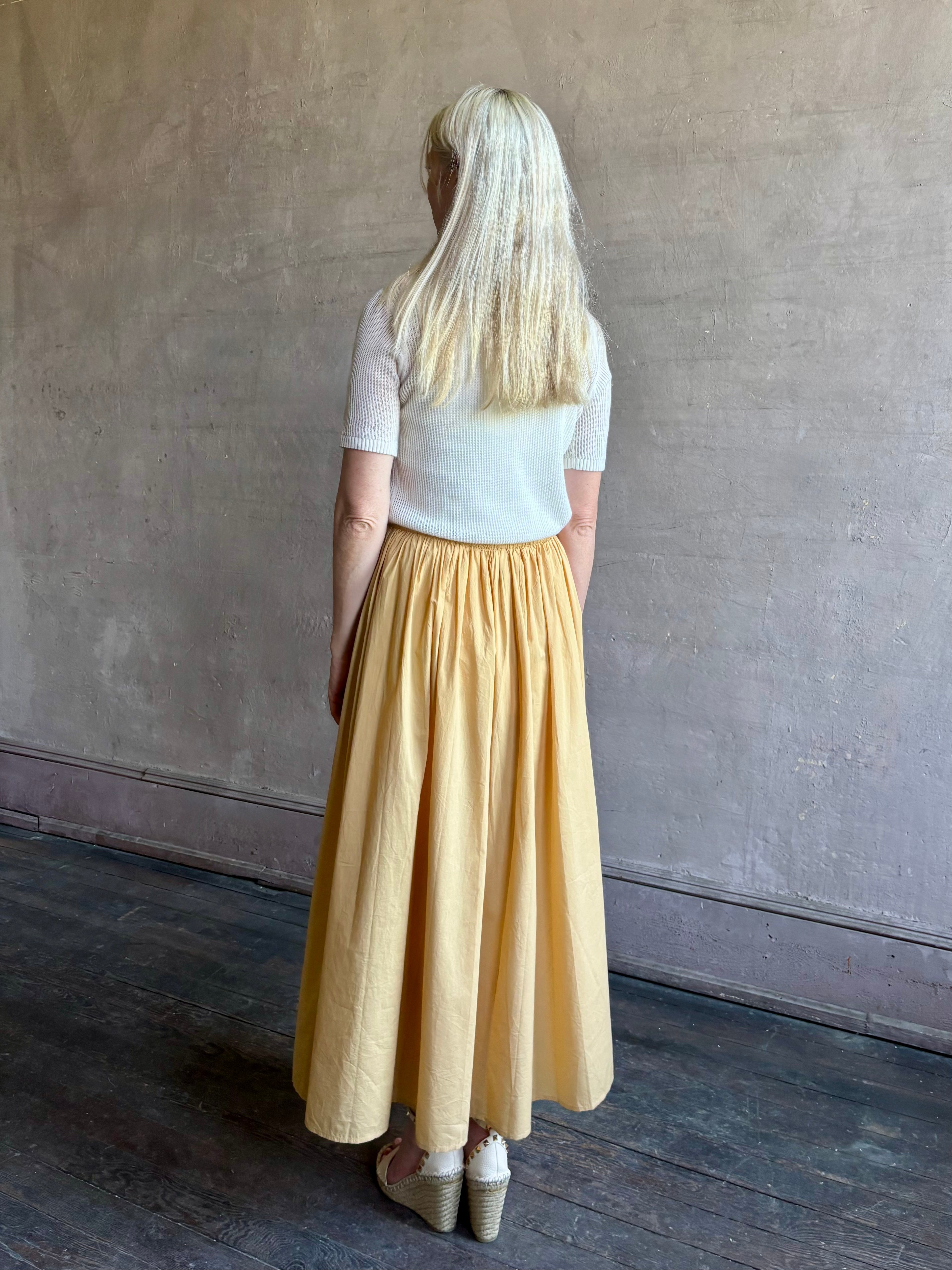 Mature woman wearing the Hanne Tie Maxi Skirt from Apiece Apart rendered in a butter yellow hue and crafted from 100% organic cotton with adjustable drawstring and elastic waistband
