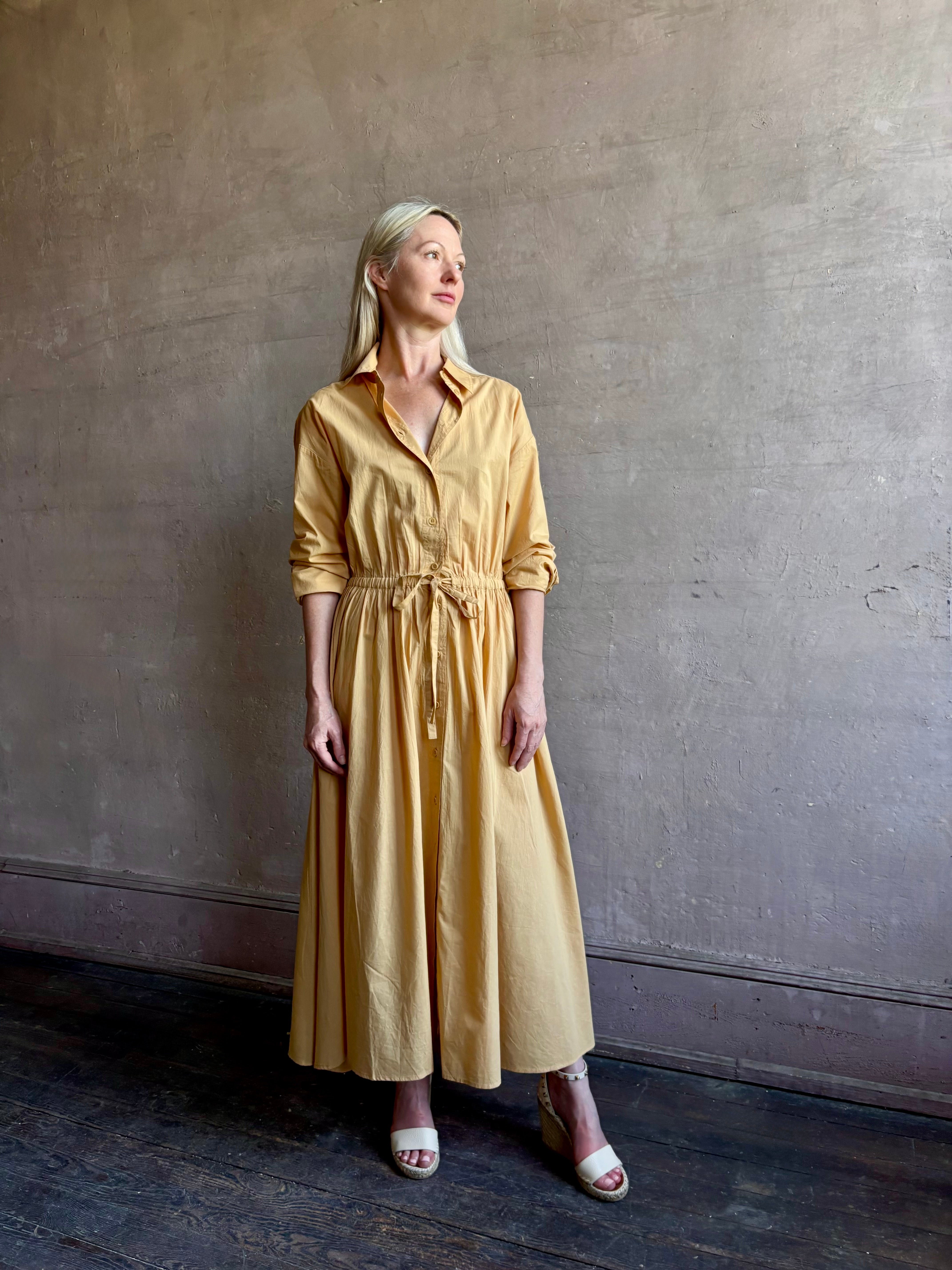 Image of mature woman wearing the Priska Shirt Dress from Apiece Apart rendered in a butter yellow and crafted from 100% organic cotton featuring long sleeves, collared button front and adjustable drawstring waistband