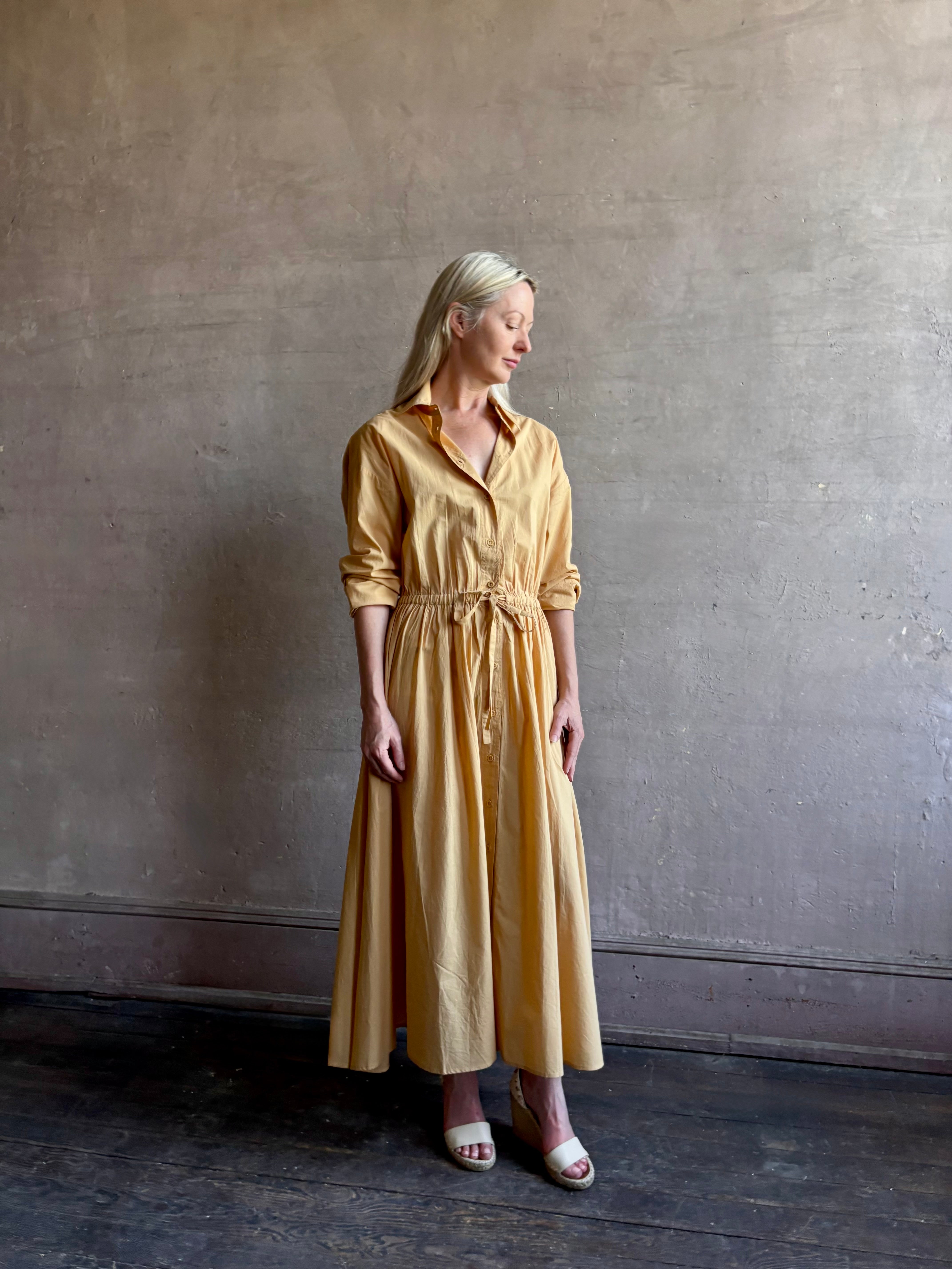 Image of mature woman wearing the Priska Shirt Dress from Apiece Apart rendered in a butter yellow and crafted from 100% organic cotton featuring long sleeves, collared button front and adjustable drawstring waistband