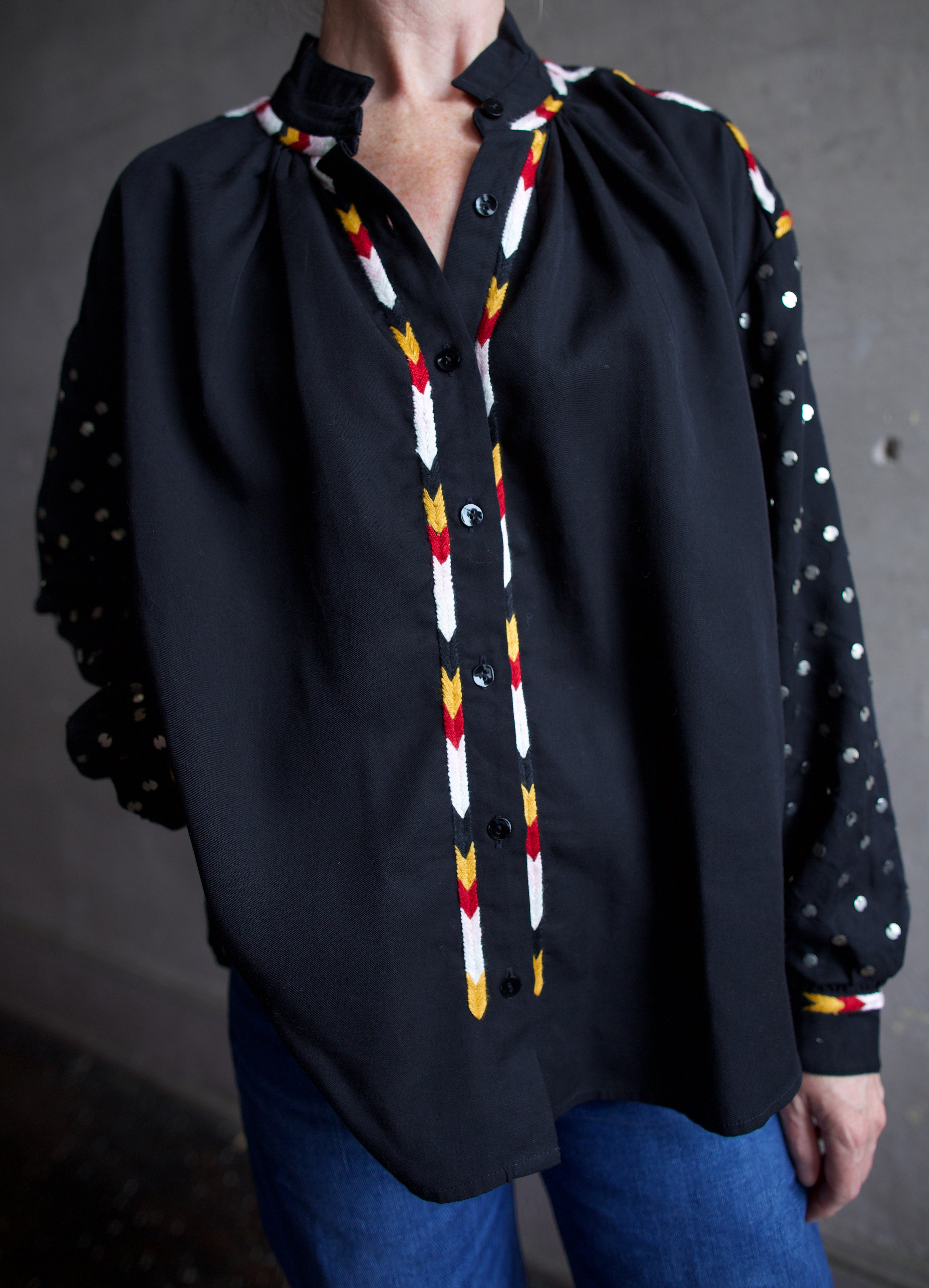 Image featuring a woman wearing the Alix of Bohemia Kiki Stardust button front black shirt with red, white, yellow, & black embroidery and metal discs on the  long sleeves.