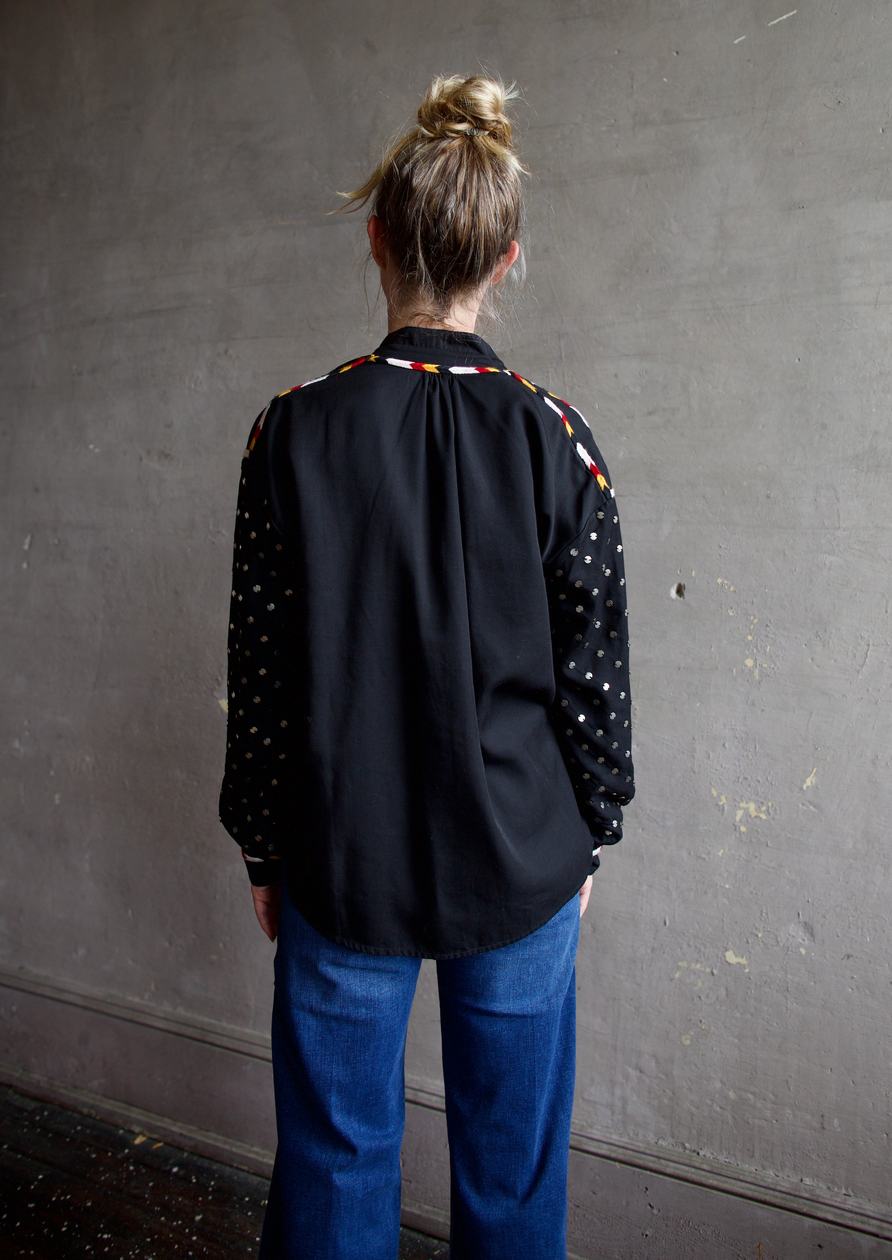 Image featuring a woman wearing the Alix of Bohemia Kiki Stardust button front black shirt with red, white, yellow, & black embroidery and metal discs on the  long sleeves.