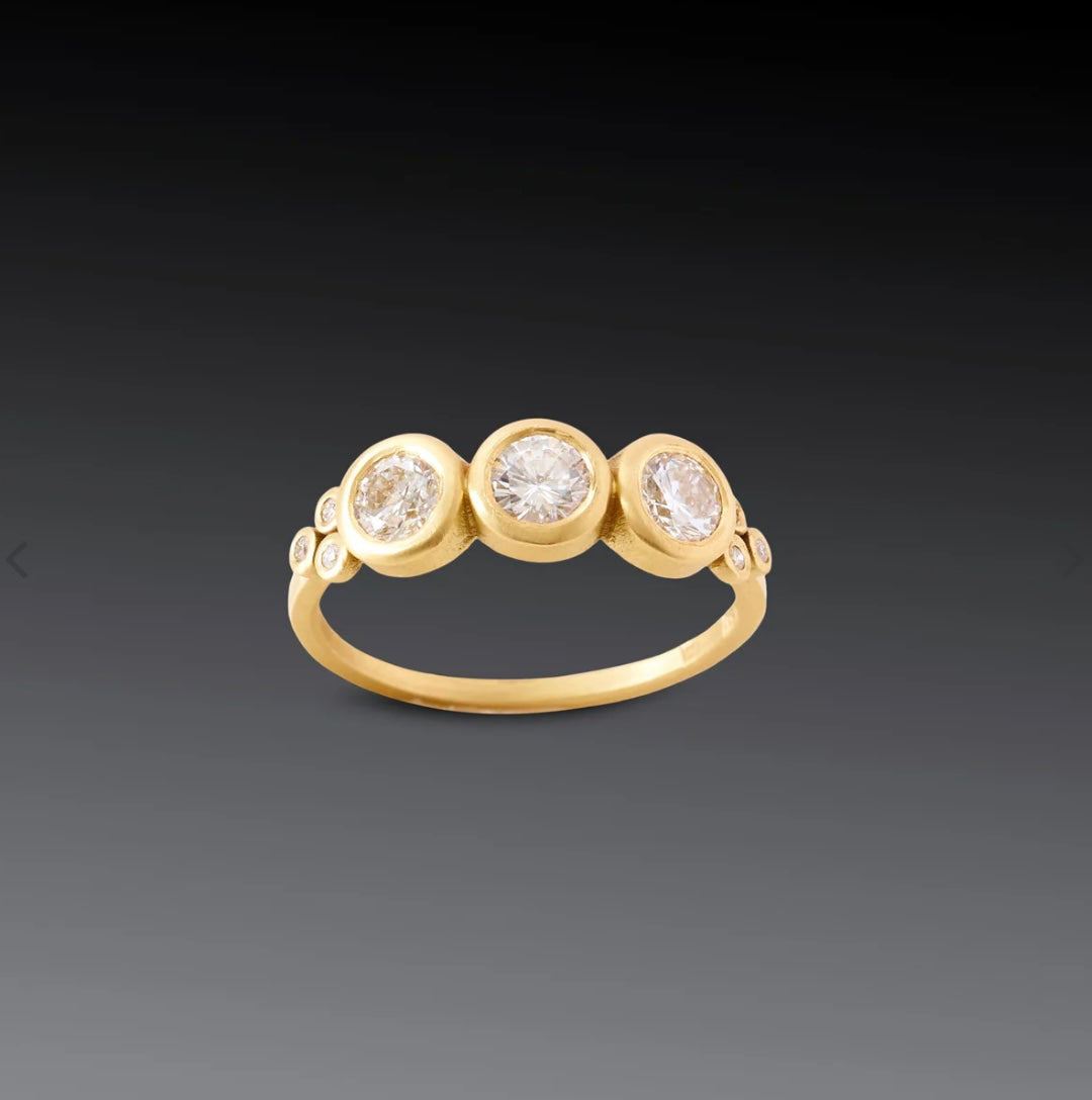 Image of a ring that features three 4mm, brilliant cut white diamonds, framed by sparkling diamond trios. The diamonds are set on a hammered 18k gold band.