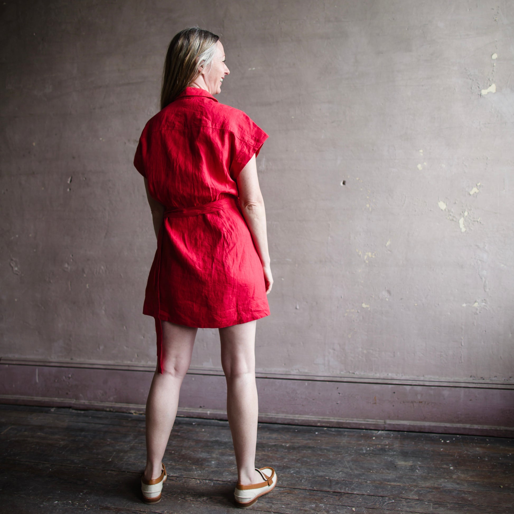 Image featuring a woman wearing the red Catania classic shirting dress from Apiece Apart with a self tie waist, relaxed bodice, dolman sleeve, shirt collar neckline and button placket.