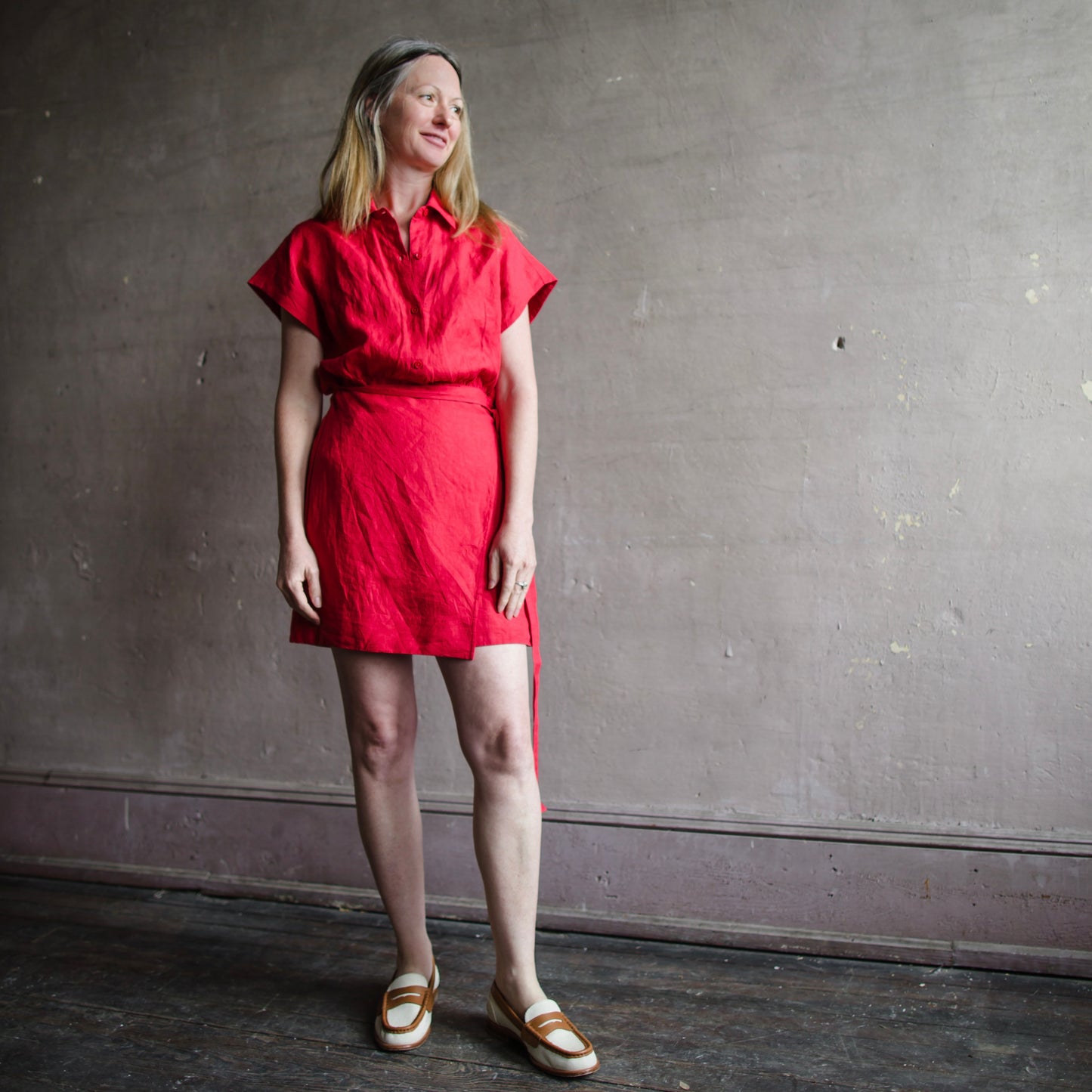 Image featuring a woman wearing the red Catania classic shirting dress from Apiece Apart with a self tie waist, relaxed bodice, dolman sleeve, shirt collar neckline and button placket.