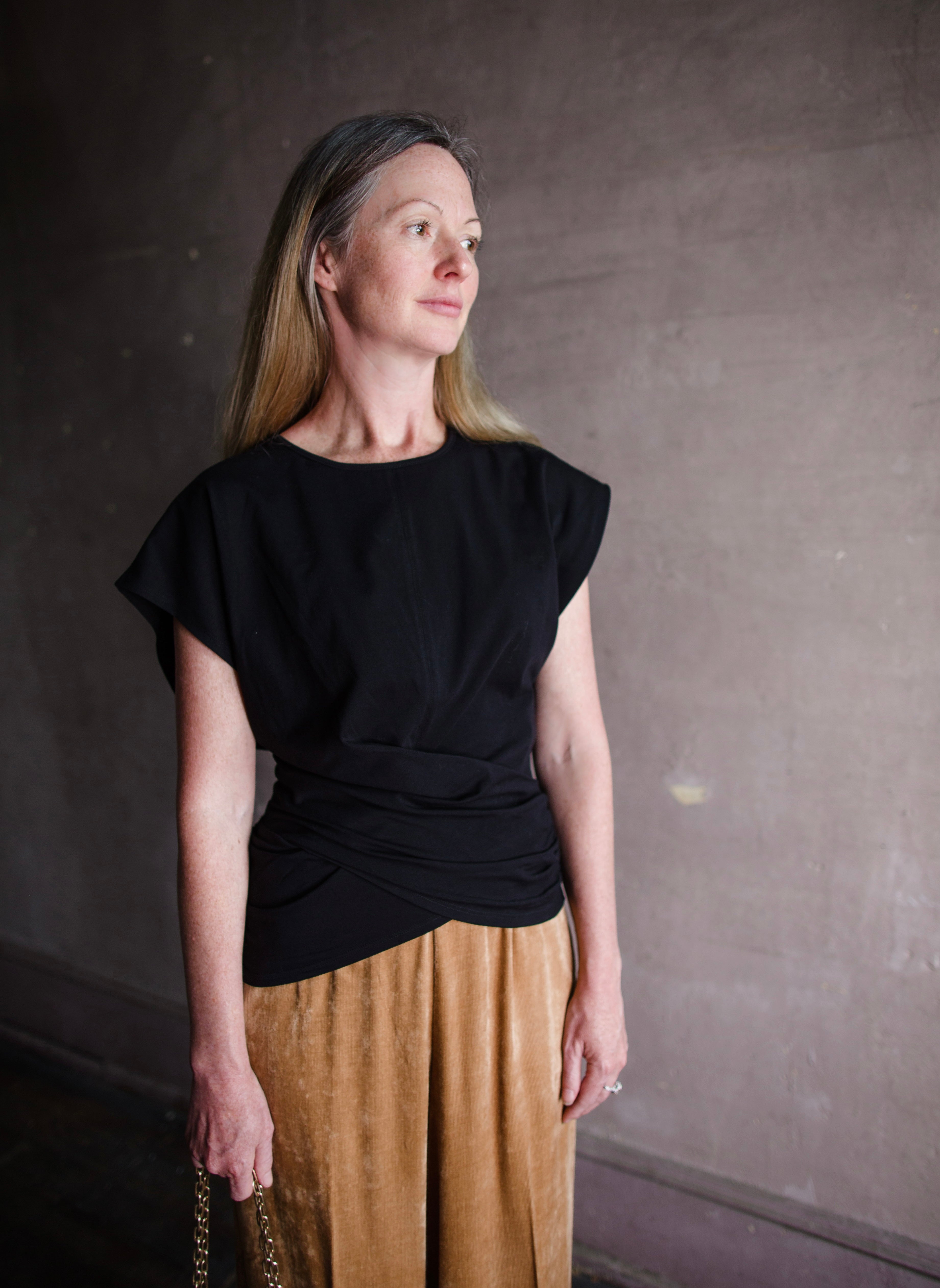 Image featuring a woman wearing the twist top by Apiece Apart with a crew neckline,  cap sleeve and a flattering twist at the waist in black.