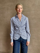 Mature woman wearing the European tailored Sierra Shirt from Callas rendered in a crisp navy and white stripe and crafted from cotton poplin featuring a flattering ruched front , long sleeves, v-neck and rounded bottom detail
