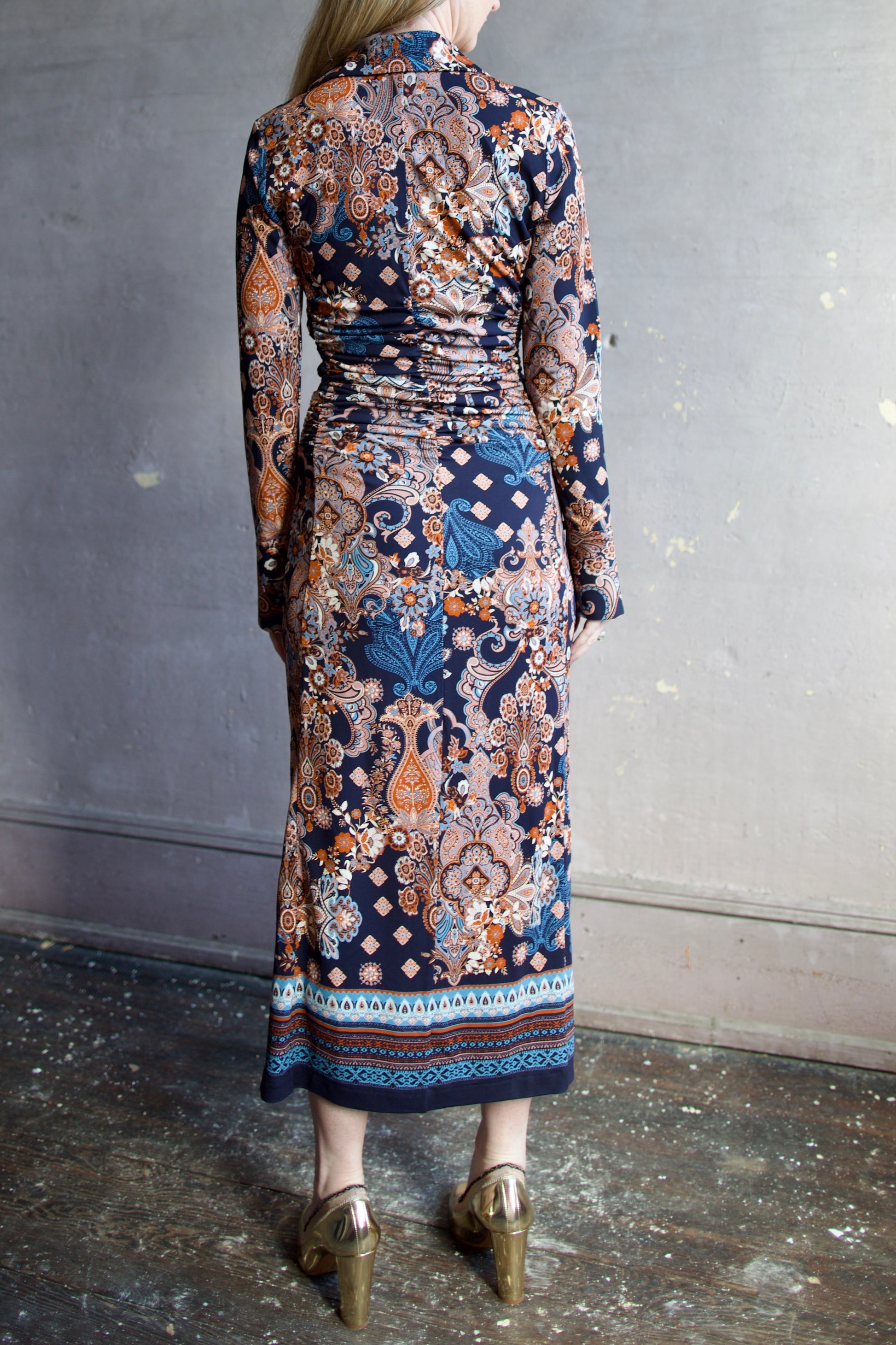 Image featuring a woman wearing the Shirley dress by Cara Cara with a button front, ruched body and side slits in a tailored fit paisley print in blues, orange, and cream.