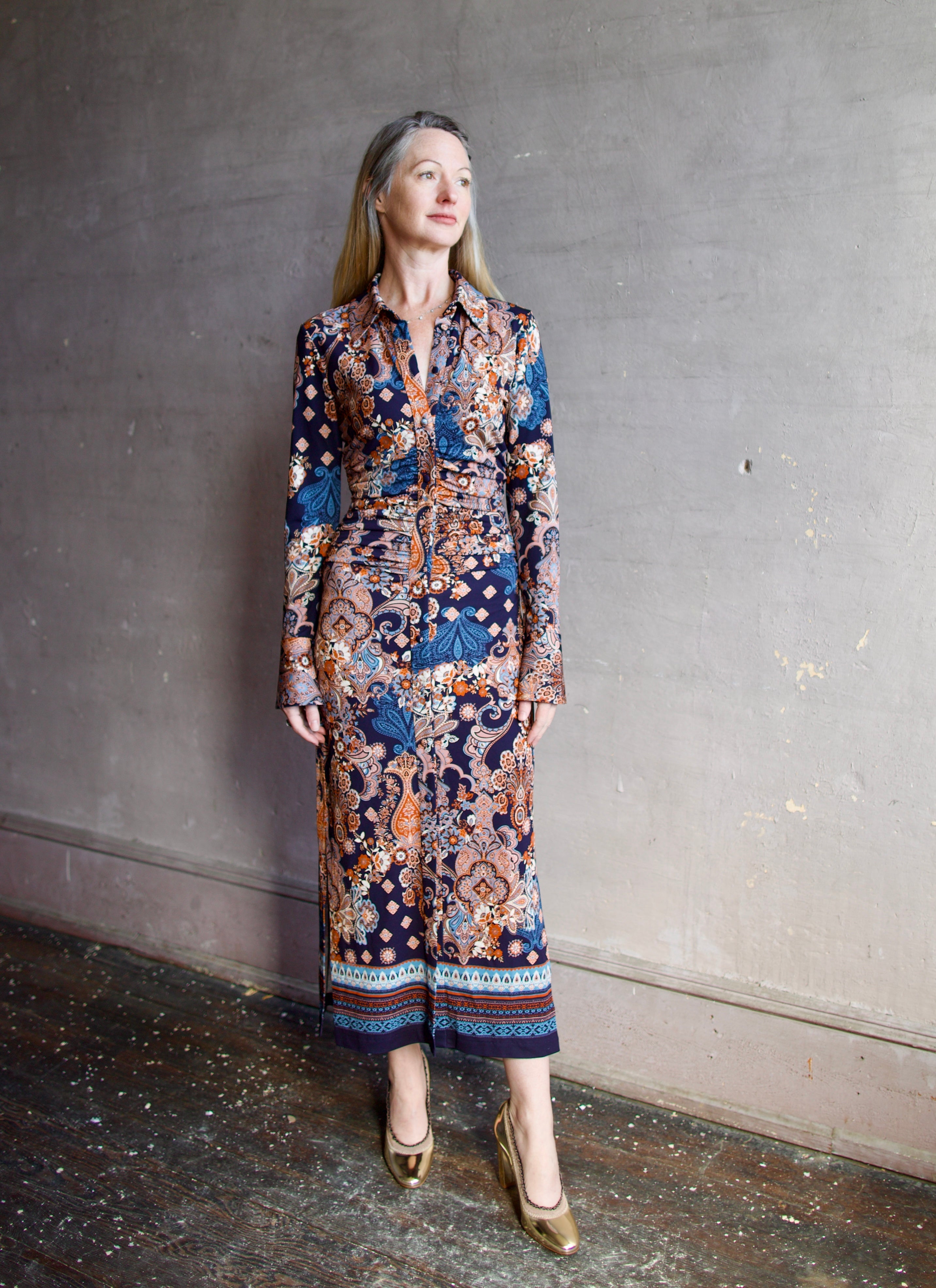 Image featuring a woman wearing the Shirley dress by Cara Cara with a button front, ruched body and side slits in a tailored fit paisley print in blues, orange, and cream.