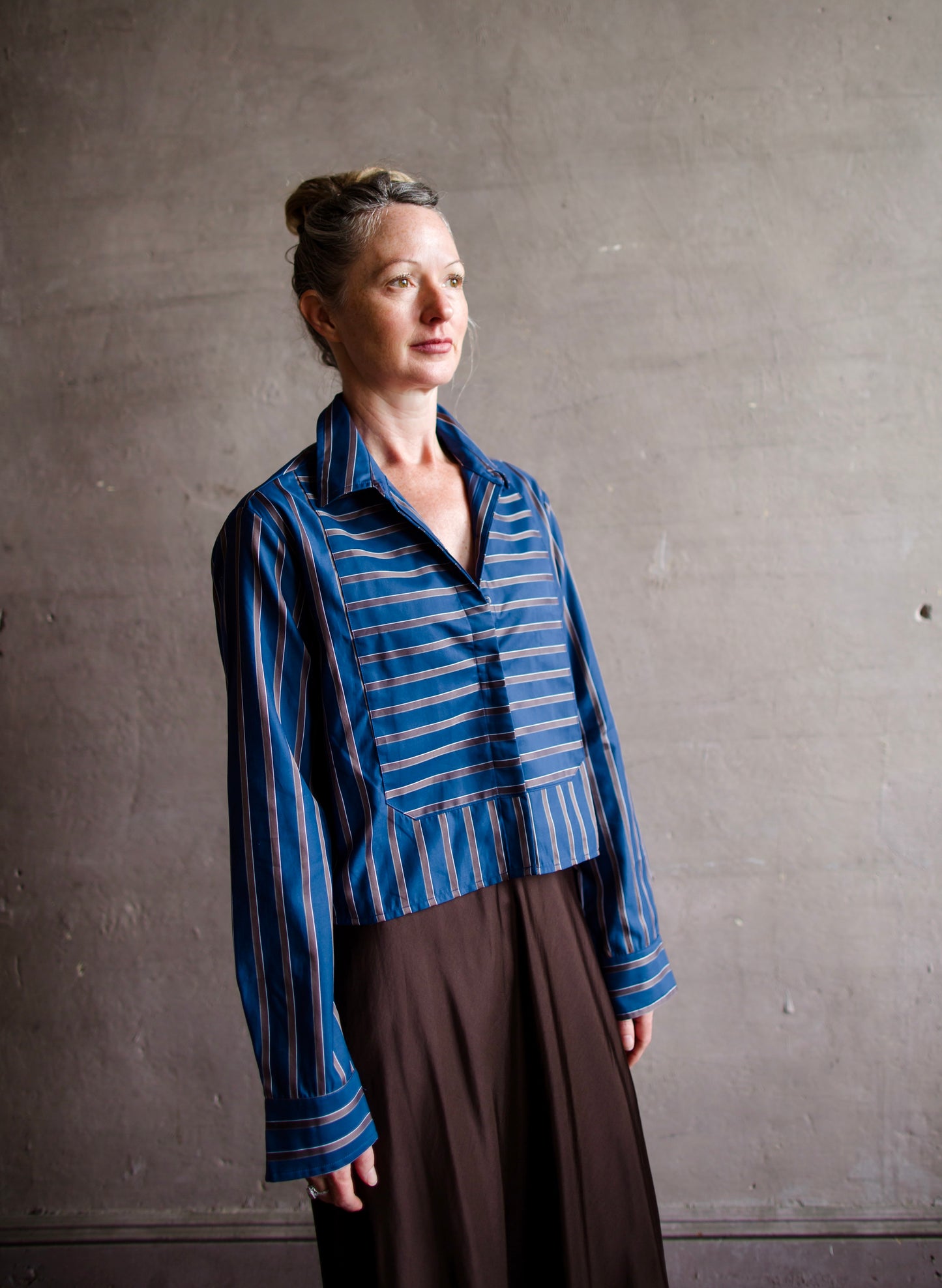 Image featuring a woman wearing the bib striped top by Cissa with a button front stripe placket and cropped, boxy fit in a blue and brown stripe.