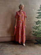 Mature woman wearing Martine maxi Dress from Trovata rendered in the Botanic Tile pink and orange floral print and crafted from a luxurious blend of cotton and silk with button front, collar, short sleeves and matching optional self tile belt