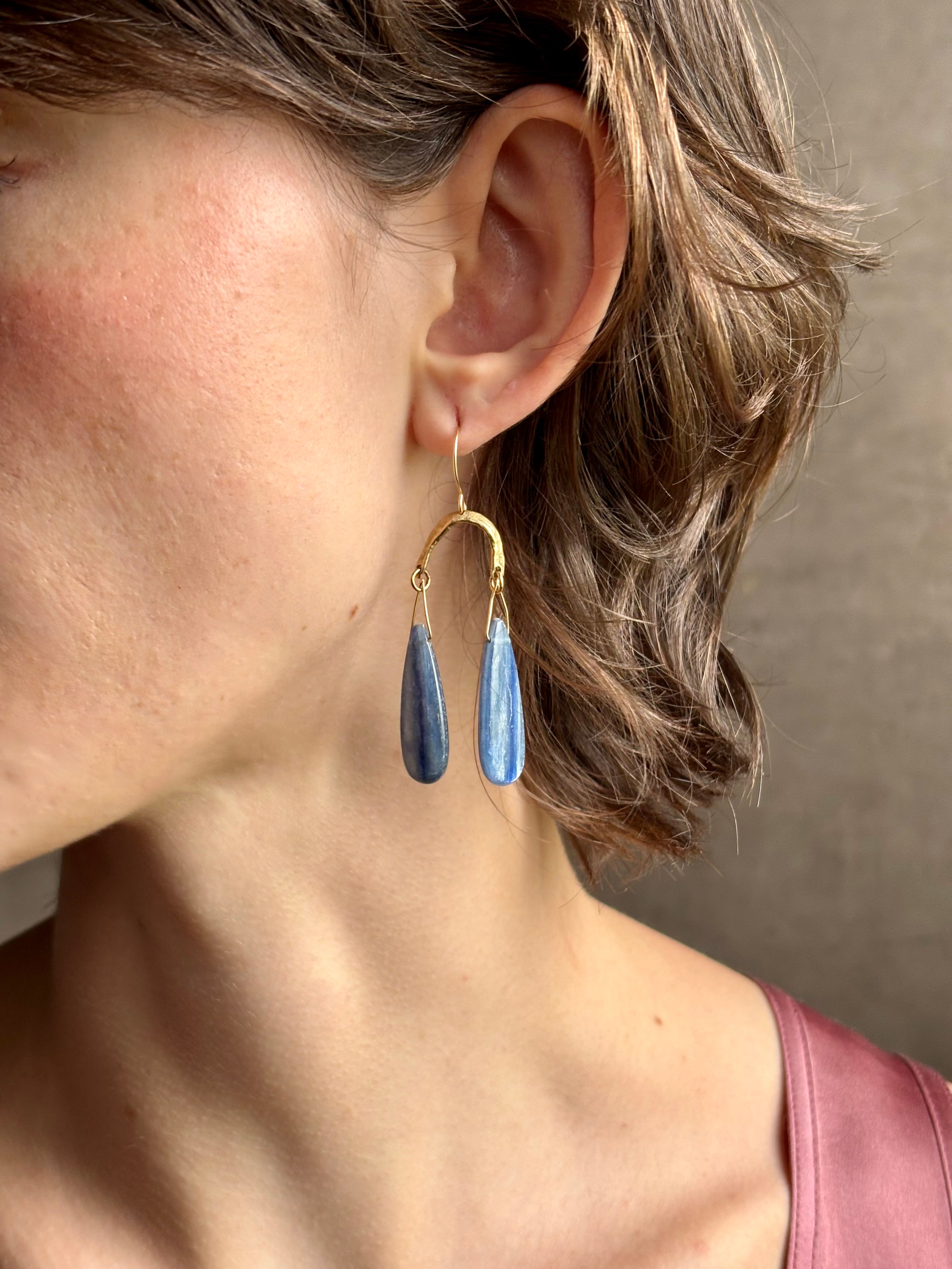 Woman wearing Delta Earrings from New York fine jewelry artist Emilie Shapiro with dangling teardrop shaped polished kyanite and warm textured half circle brass dangle gracefully from handmade gold filled earring wires
