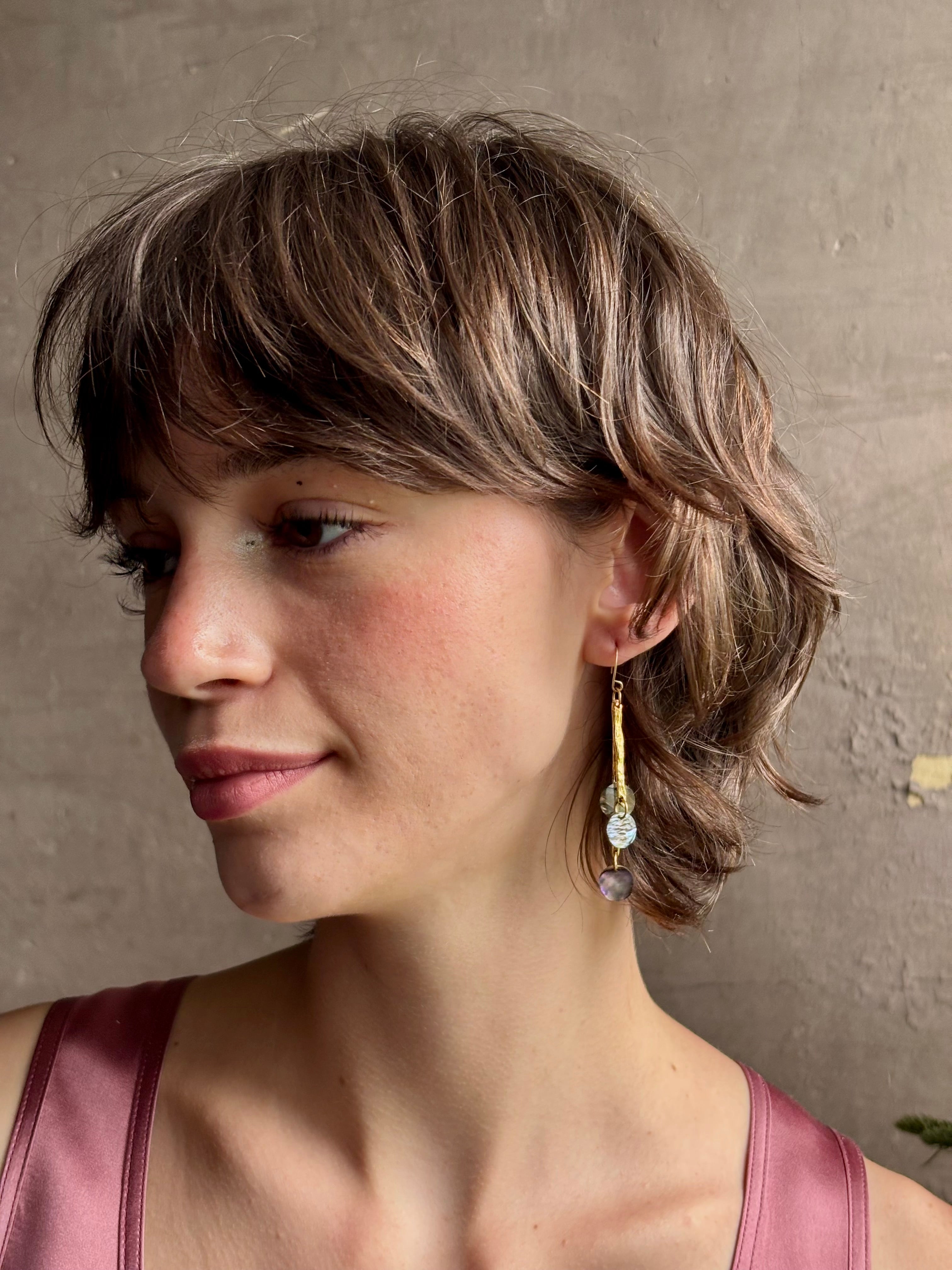 Woman wearing Lyra Earrings from designer jewelry artist Emilie Shapiro with iridescent paua shells dangling from textured brass and a gold filled wire hook with beautiful movement handcrafted in USA