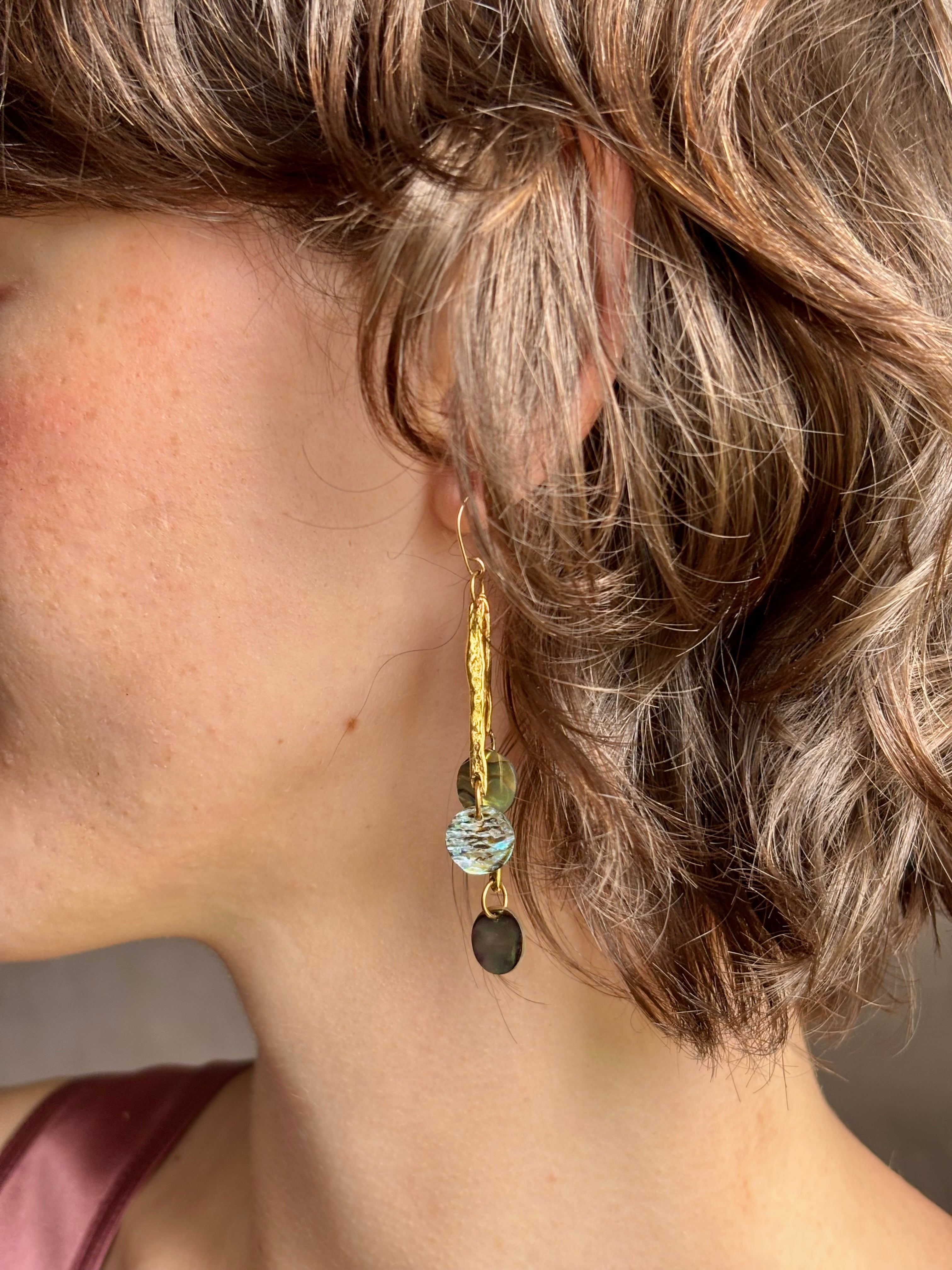 Woman wearing Lyra Earrings from designer jewelry artist Emilie Shapiro with iridescent paua shells dangling from textured brass and a gold filled wire hook with beautiful movement handcrafted in USA