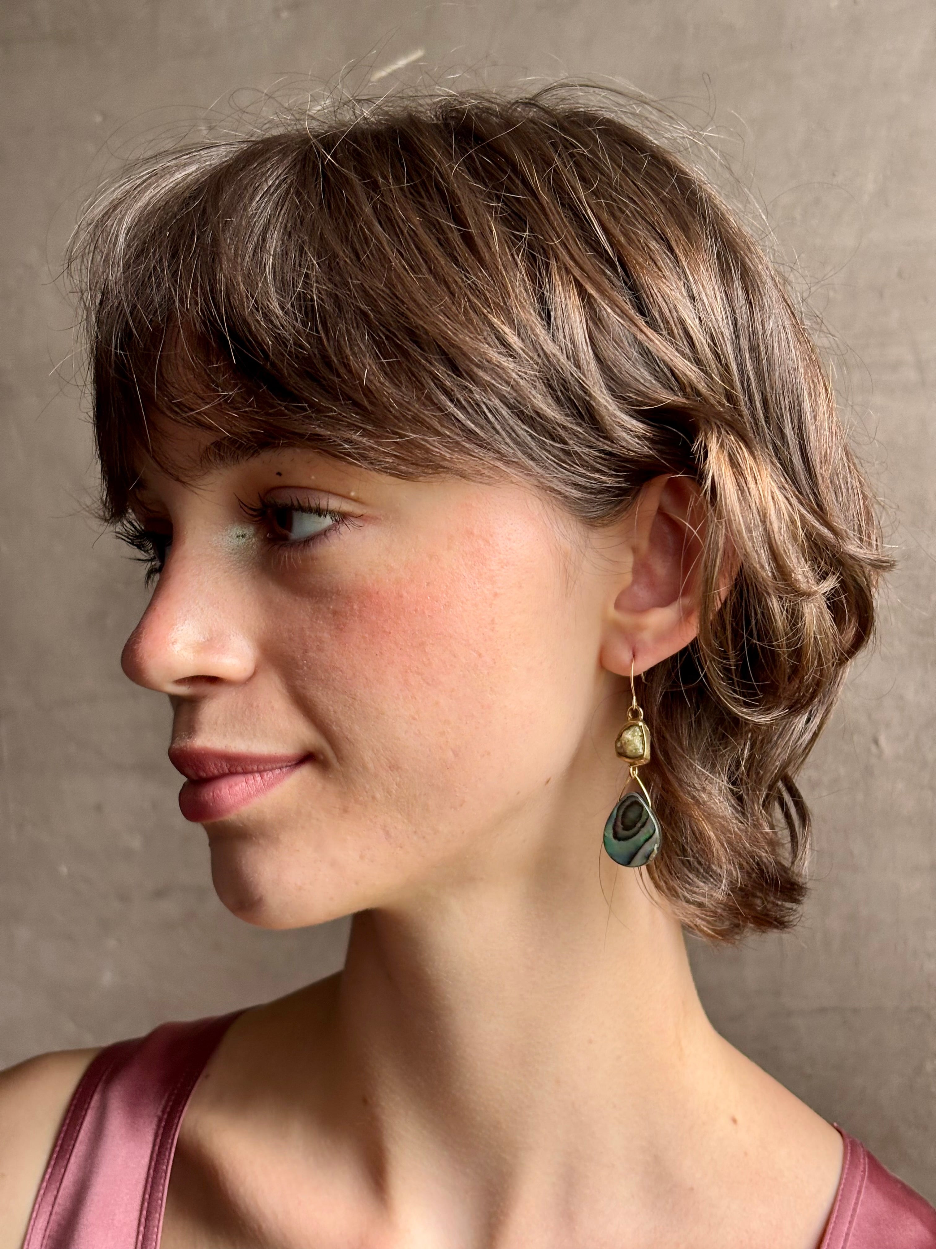 Woman wearing Northern Light Earrings from jewelry artist Emilie Shapiro with a tear shaped paua shell dangling from a natural garnet mounted in brass and suspended from a gold filled hook handcrafted in USA
