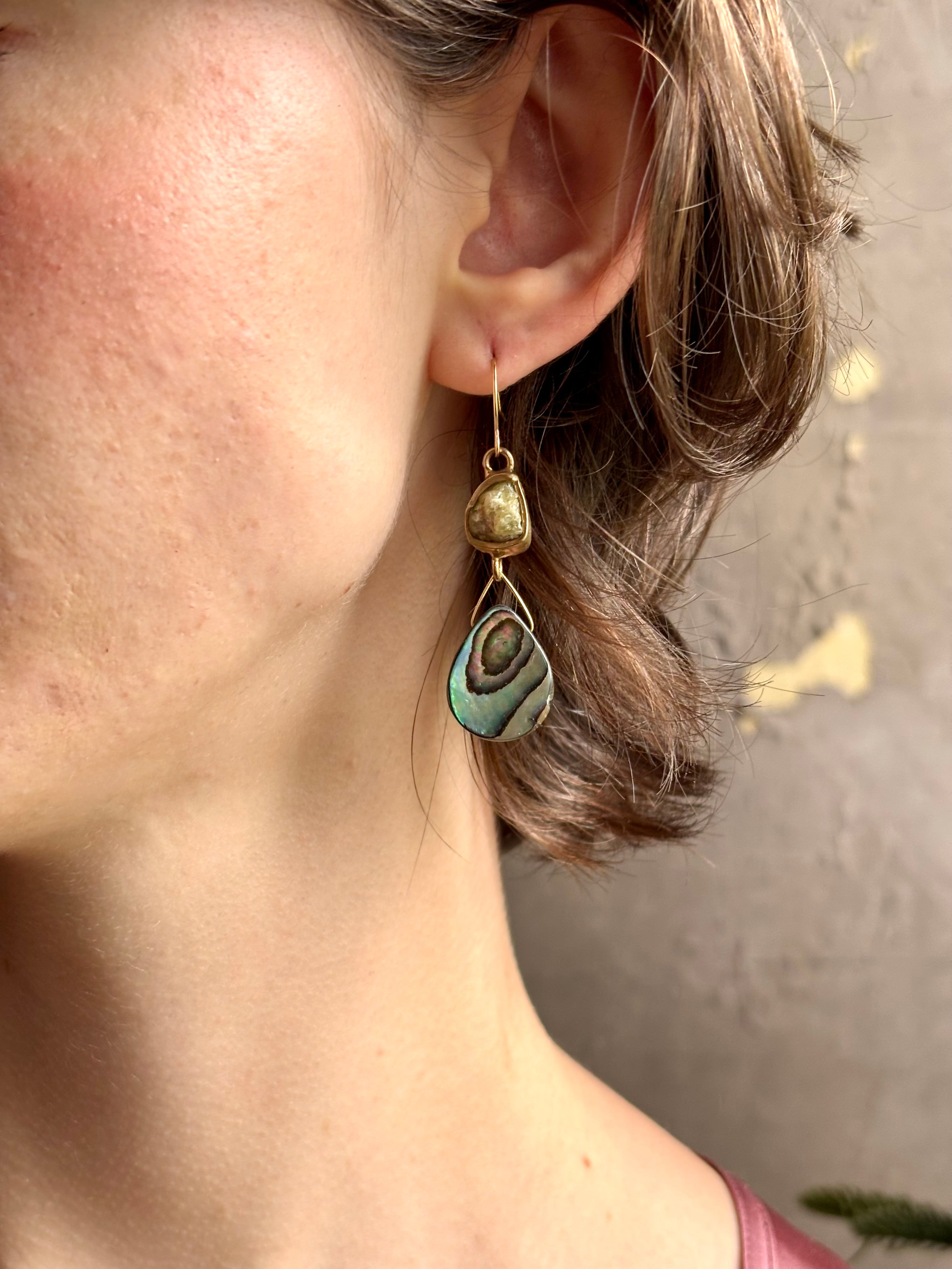 Woman wearing Northern Light Earrings from jewelry artist Emilie Shapiro with a tear shaped paua shell dangling from a natural garnet mounted in brass and suspended from a gold filled hook handcrafted in USA