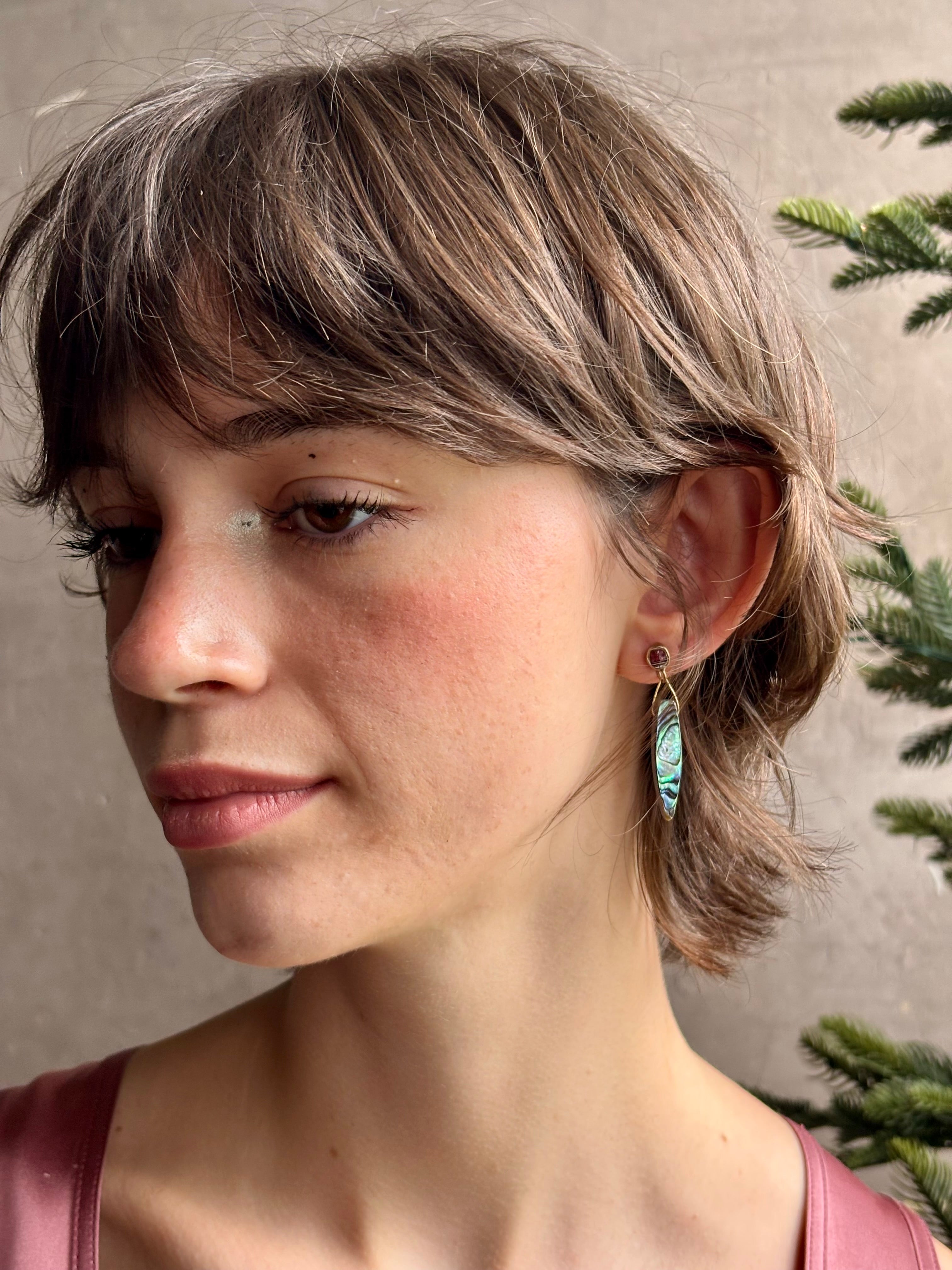 Woman wearing Umbra Earrings from fine jewelry designer Emilie Shapiro with a delicate pink spinel stud  anchoring an oval paua shell dandling from a brass triangle wire made in USA