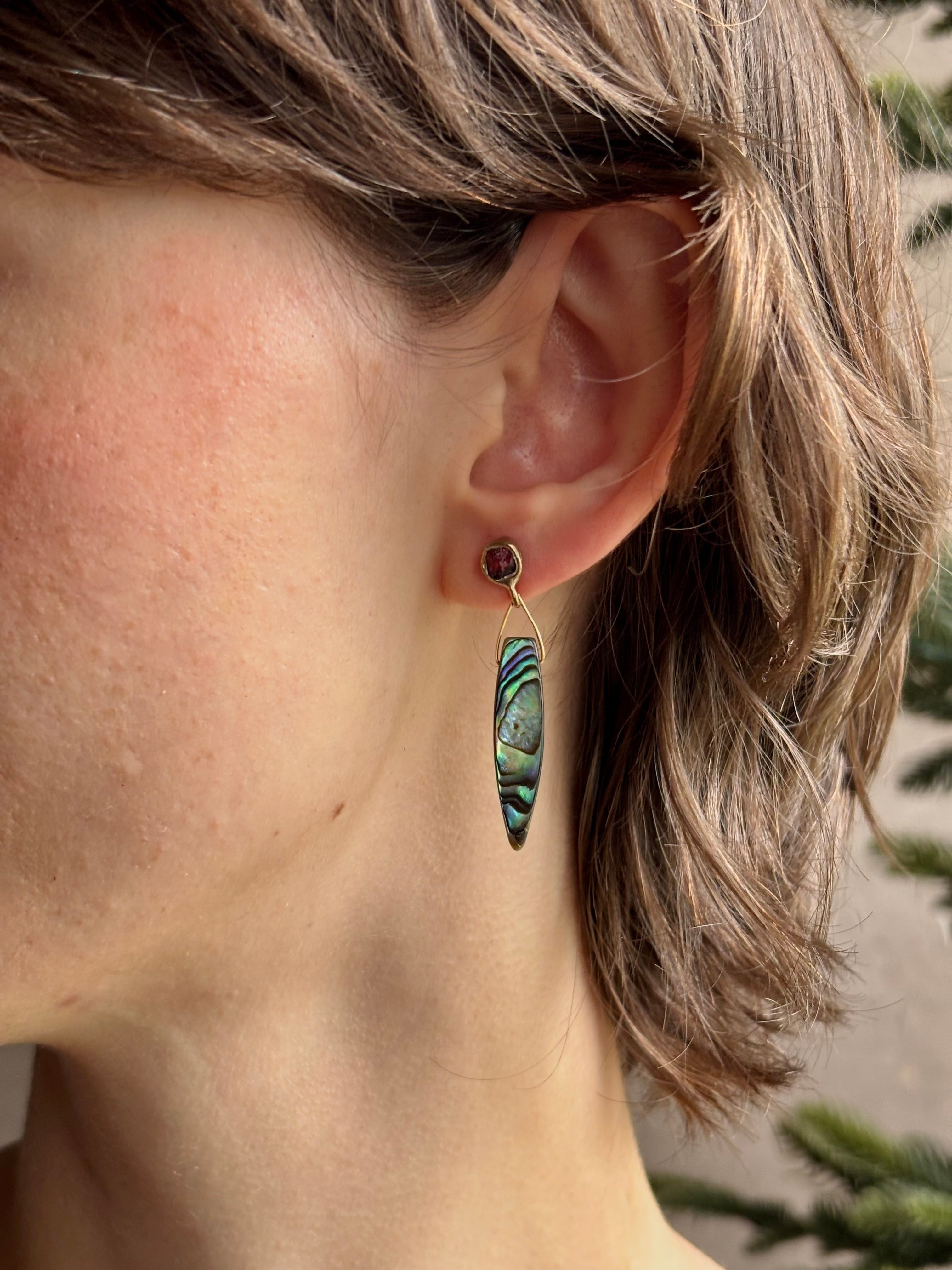 Woman wearing Umbra Earrings from fine jewelry designer Emilie Shapiro with a delicate pink spinel stud  anchoring an oval paua shell dandling from a brass triangle wire made in USA