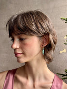 Woman wearing Umbra Earrings from fine jewelry designer Emilie Shapiro with a delicate pink spinel stud  anchoring an oval paua shell dandling from a brass triangle wire made in USA