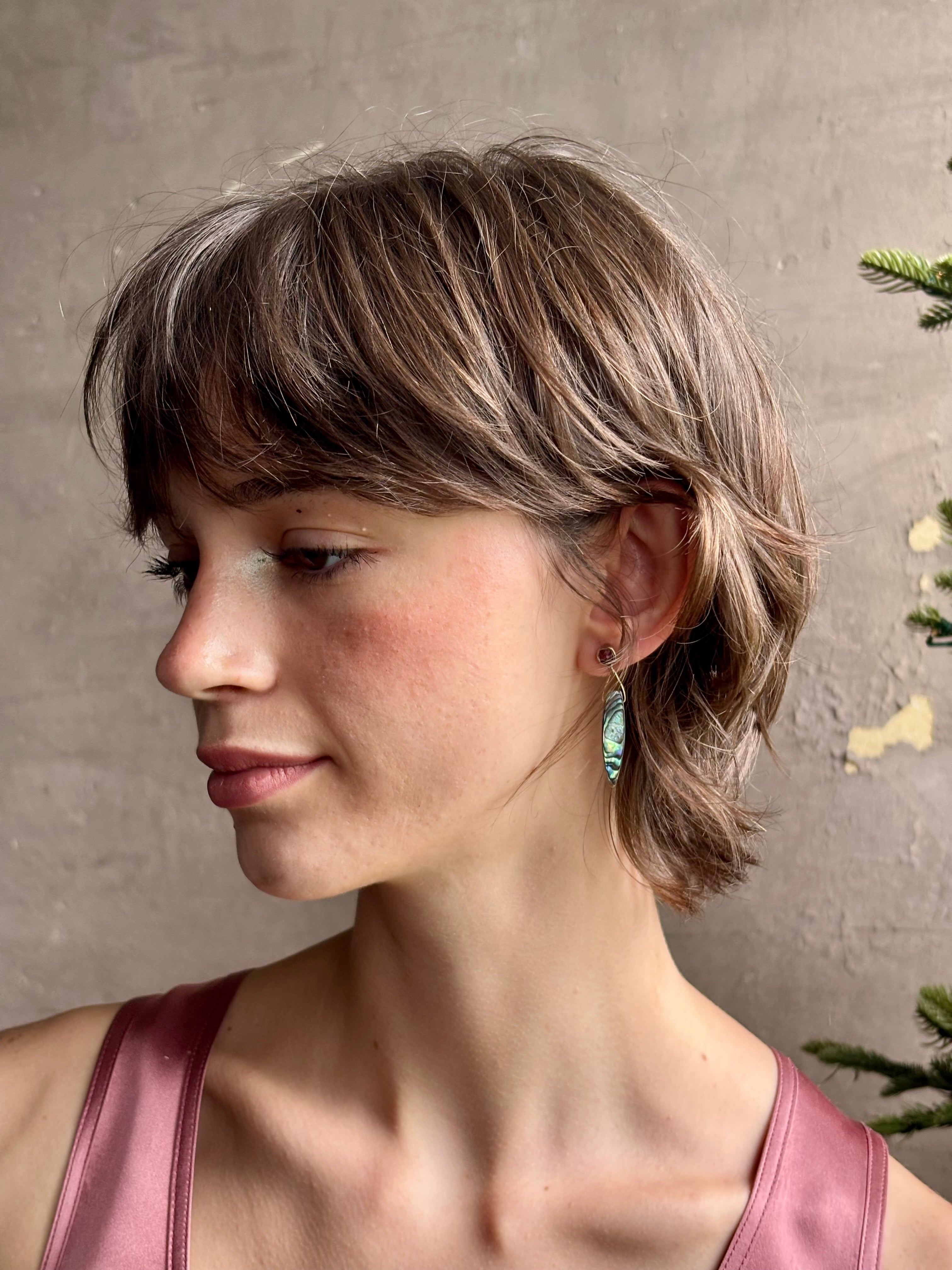 Woman wearing Umbra Earrings from fine jewelry designer Emilie Shapiro with a delicate pink spinel stud  anchoring an oval paua shell dandling from a brass triangle wire made in USA