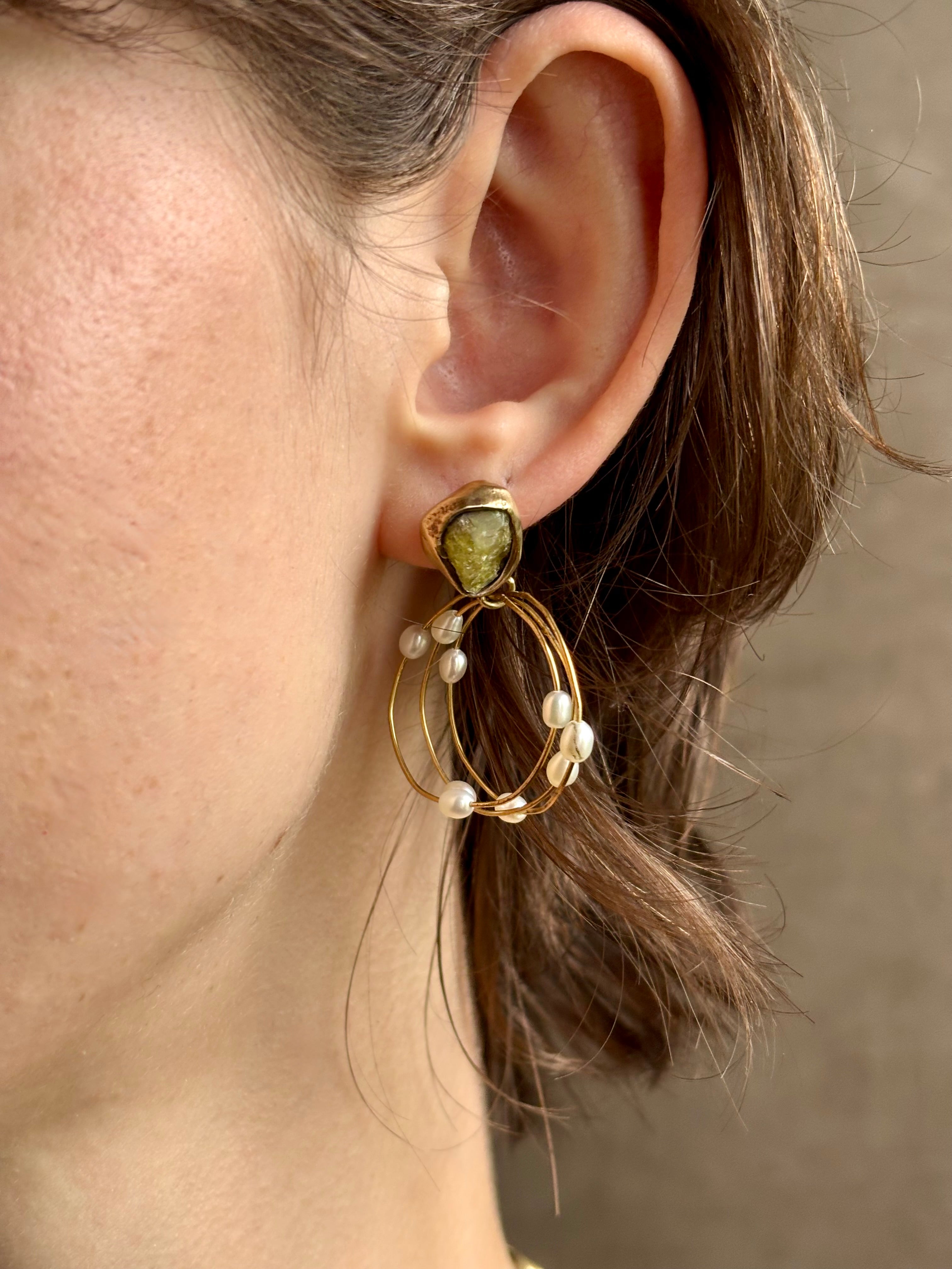 Woman wearing water lily hoop earrings from jewelry designer Emilie Shapiro with posts on Natural garnets individually embedded in warm brass that have three brass circular hoops with three natural white  rice pearls on each hoop