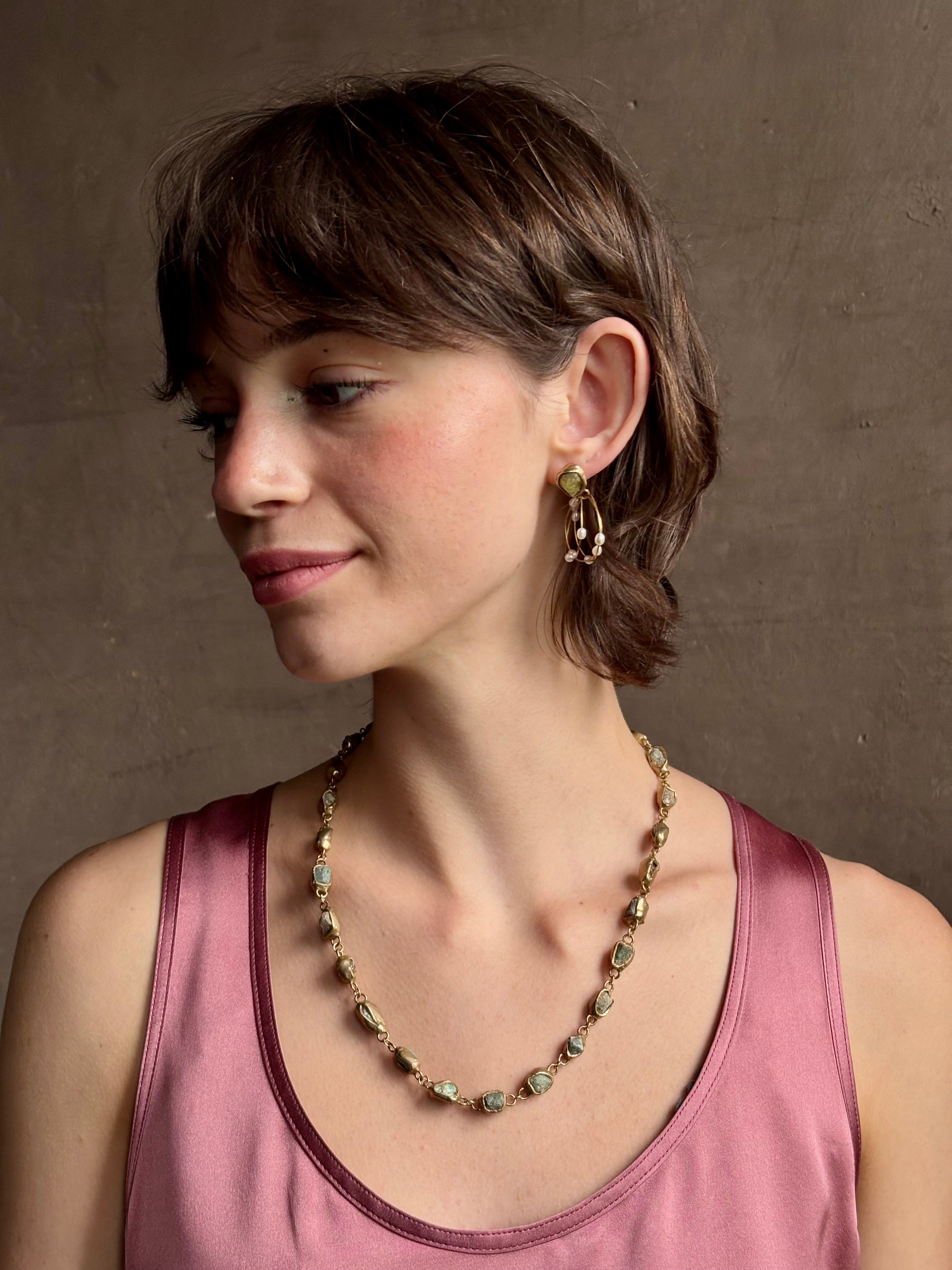 Woman wearing water lily hoop earrings from jewelry designer Emilie Shapiro with posts on Natural garnets individually embedded in warm brass that have three brass circular hoops with three natural white  rice pearls on each hoop