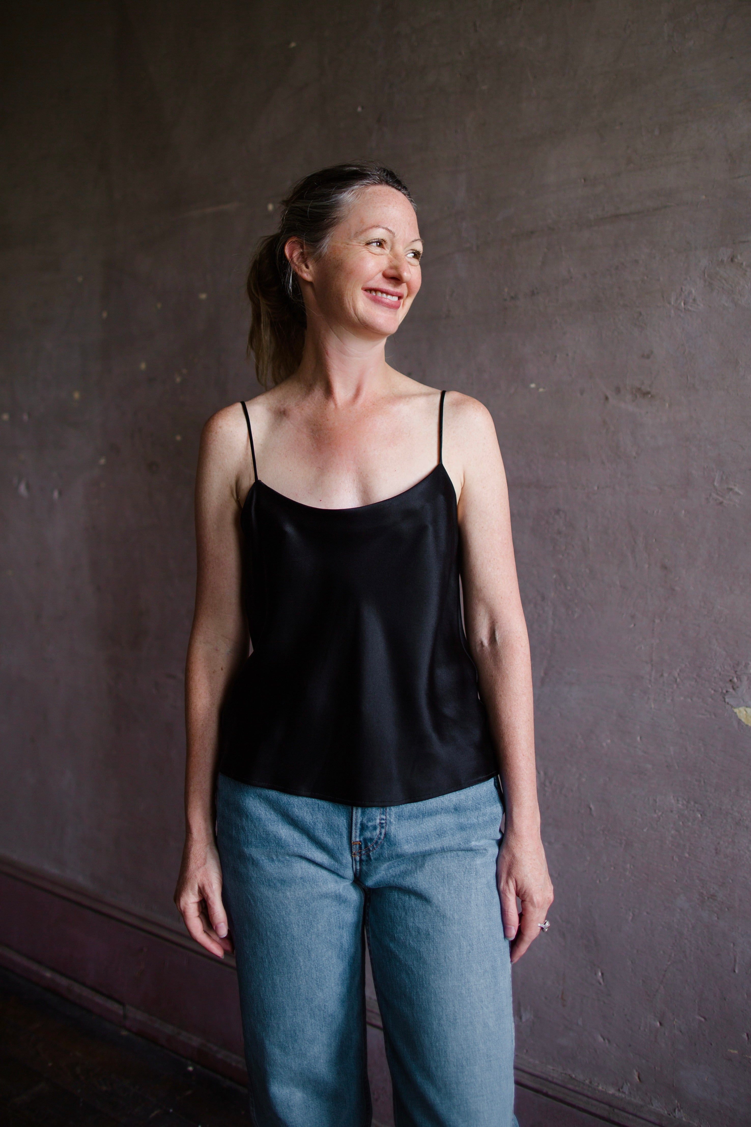 Image featuring a woman wearing the black Silk Satin Camisole from Enza Costa with a feminine scoop neckline, delicate straps, a gently flared hem and relaxed fit.