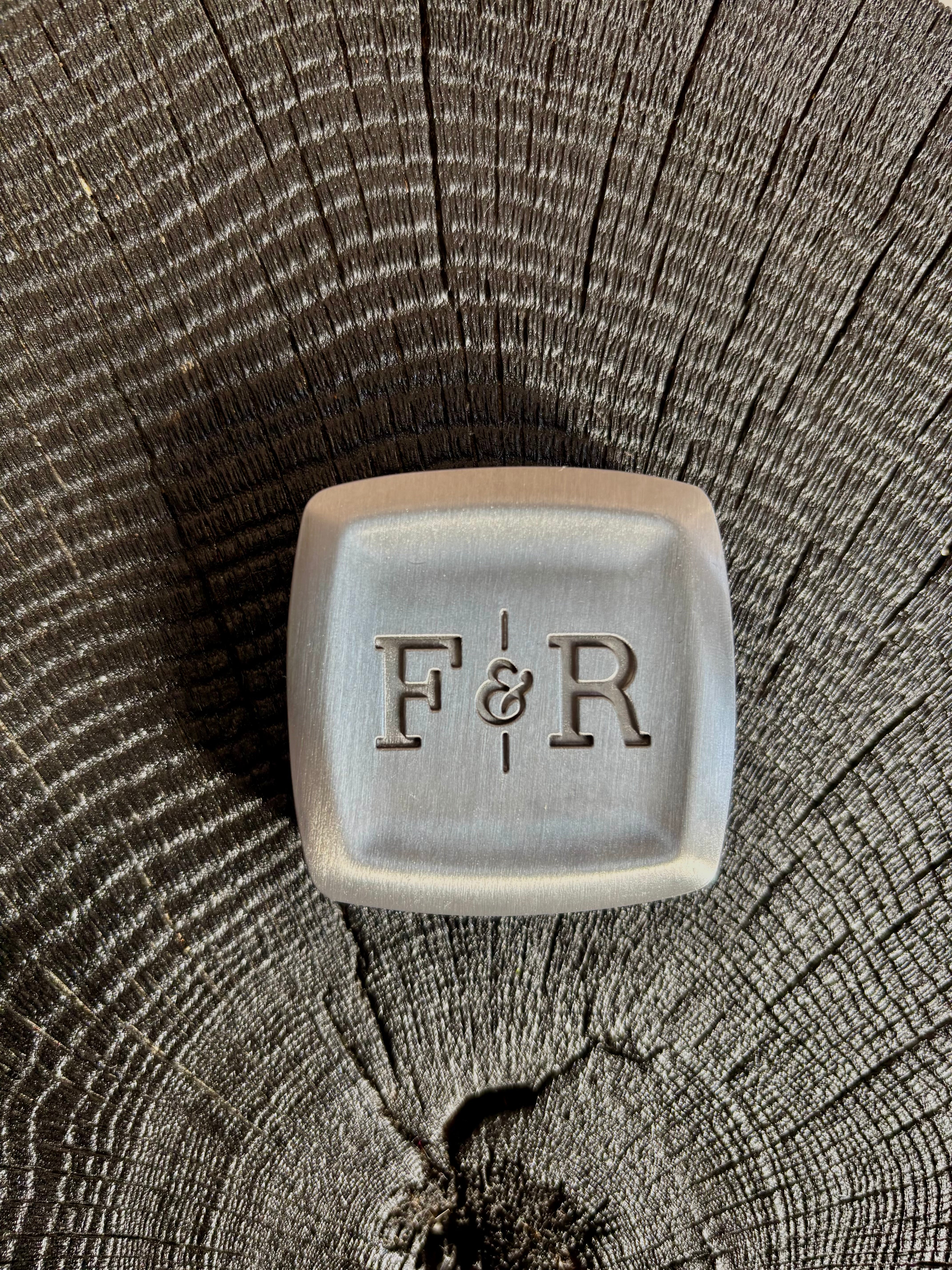 A square metal holder with rounded corners and the embossed initials 'F&R' in the center with magnet closure and refillable solid scent well