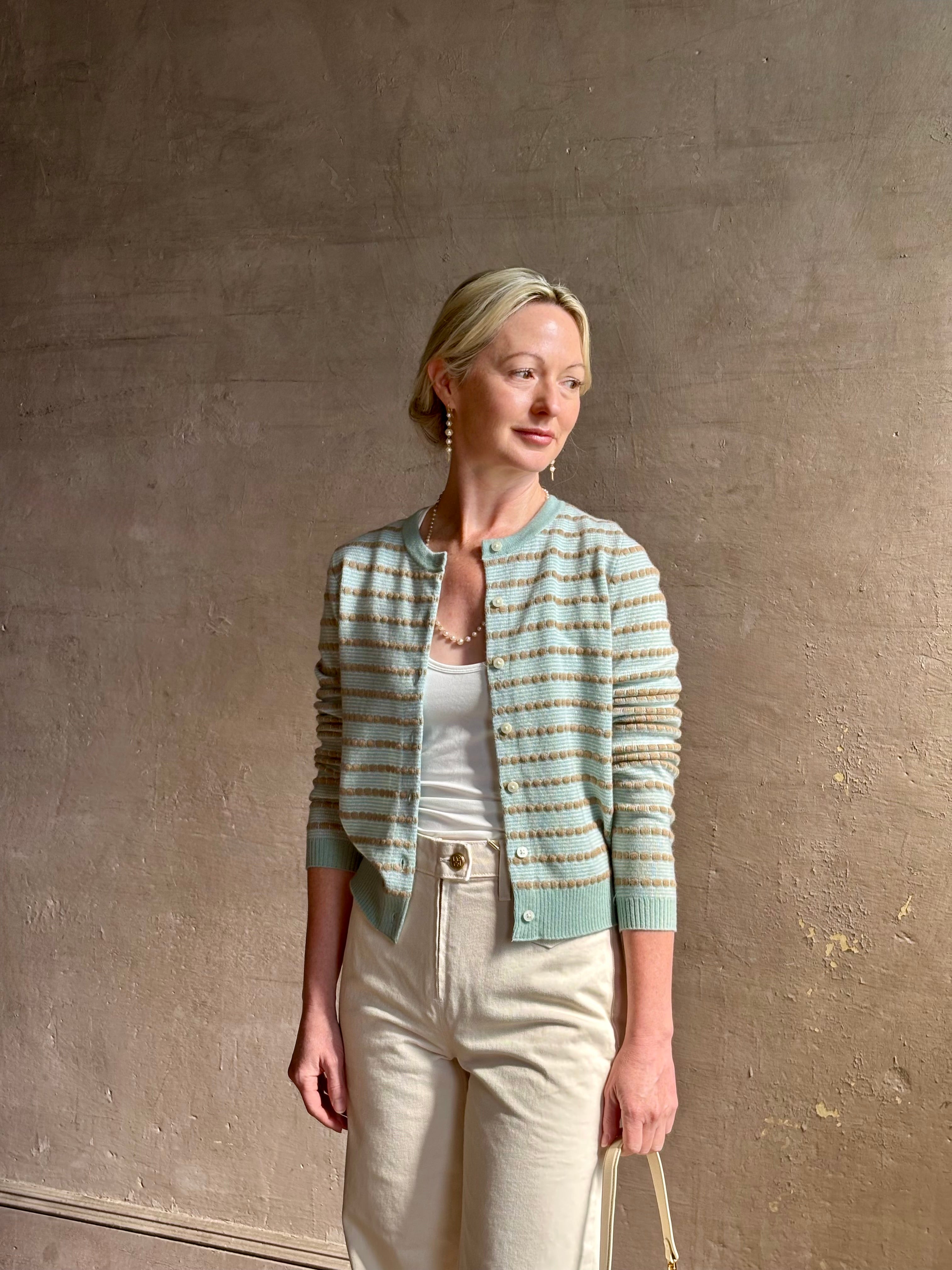 Mature woman wearing Pebble Stripe Jane Cardigan from Guest in Residence rendered in a serene sage brush aqua Khaki combo and crafted from 100% cashmere with crew neck, long sleeves and button front