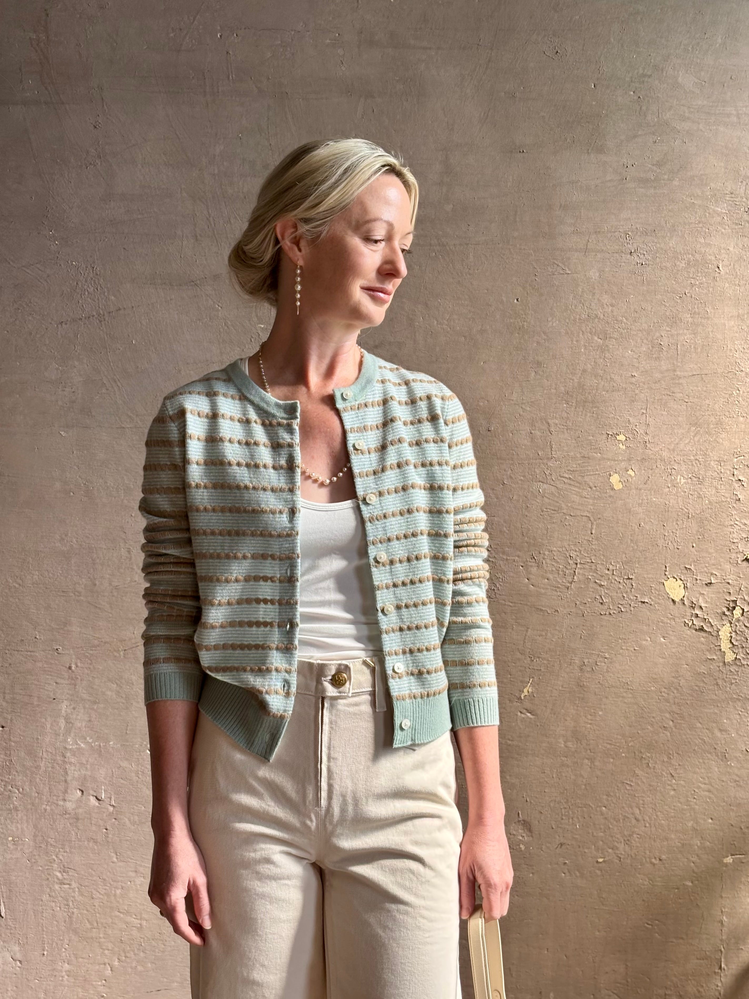 Mature woman wearing Pebble Stripe Jane Cardigan from Guest in Residence rendered in a serene sage brush aqua Khaki combo and crafted from 100% cashmere with crew neck, long sleeves and button front