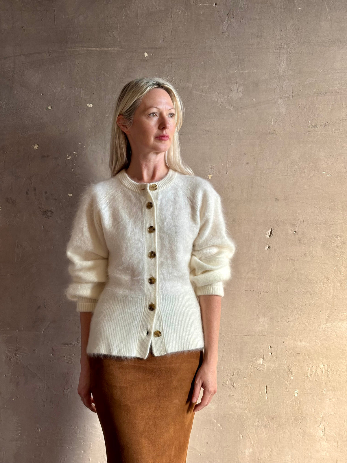 A woman wearing a cream-colored fuzzy knit cardigan with brown buttons and a brown skirt, standing against a textured beige wall.