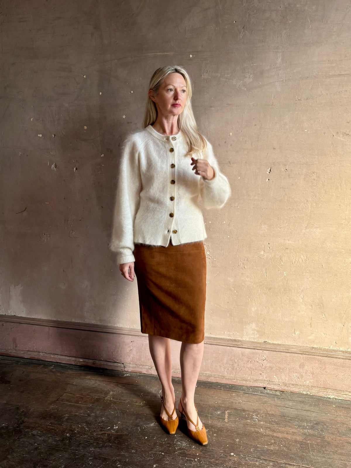 A woman stands against a textured beige wall wearing a cream-colored fuzzy button-front cardigan, a brown knee-length skirt, and Fendi Shoes