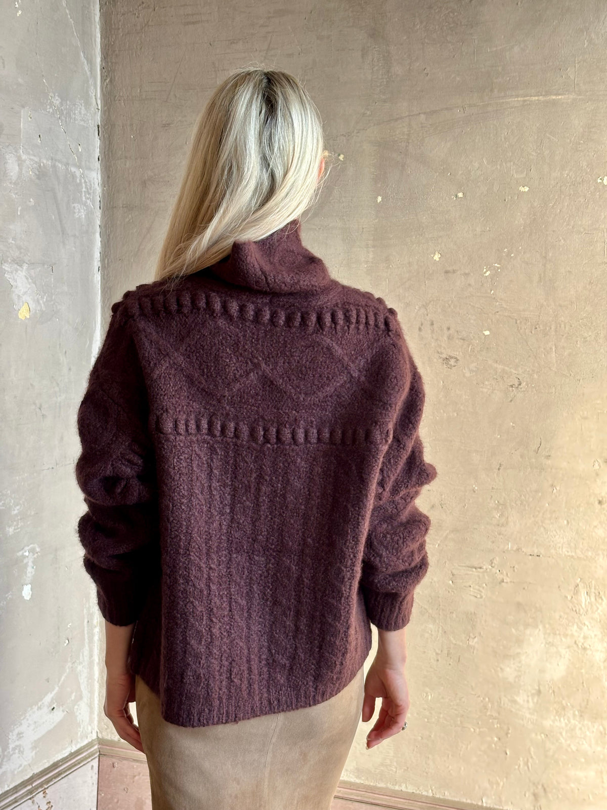 A woman shown from behind wearing a dark plum-purple textured soft cable knit cashmere silk sweater by Guest in Residence Super Cloud Turtle Neck in Fig