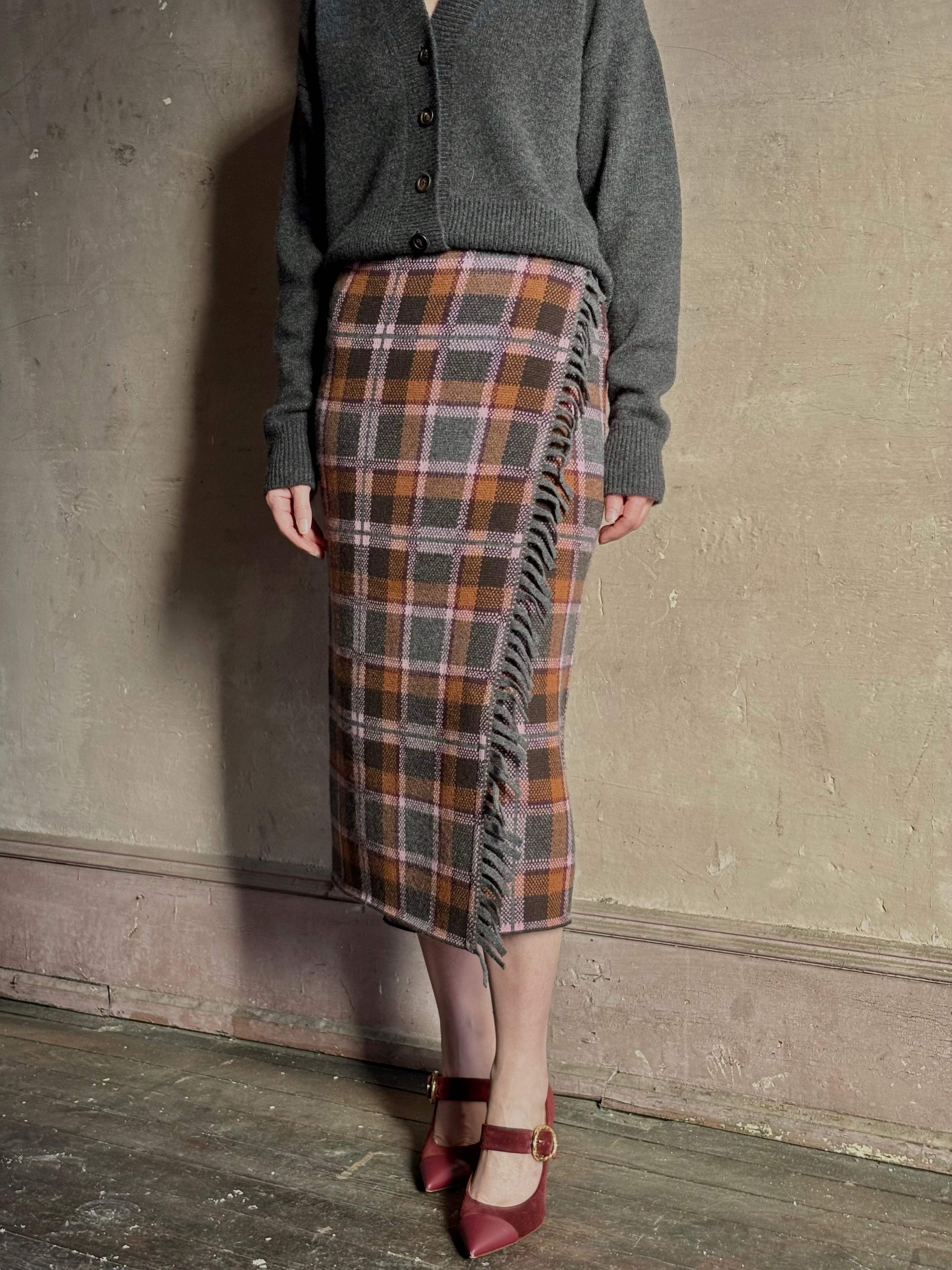 Image of mature woman wearing a gray cardigan with the Guest in Residence Tartan wrap pencil skirt in  brown, orange, and gray plaid with fringe trim 