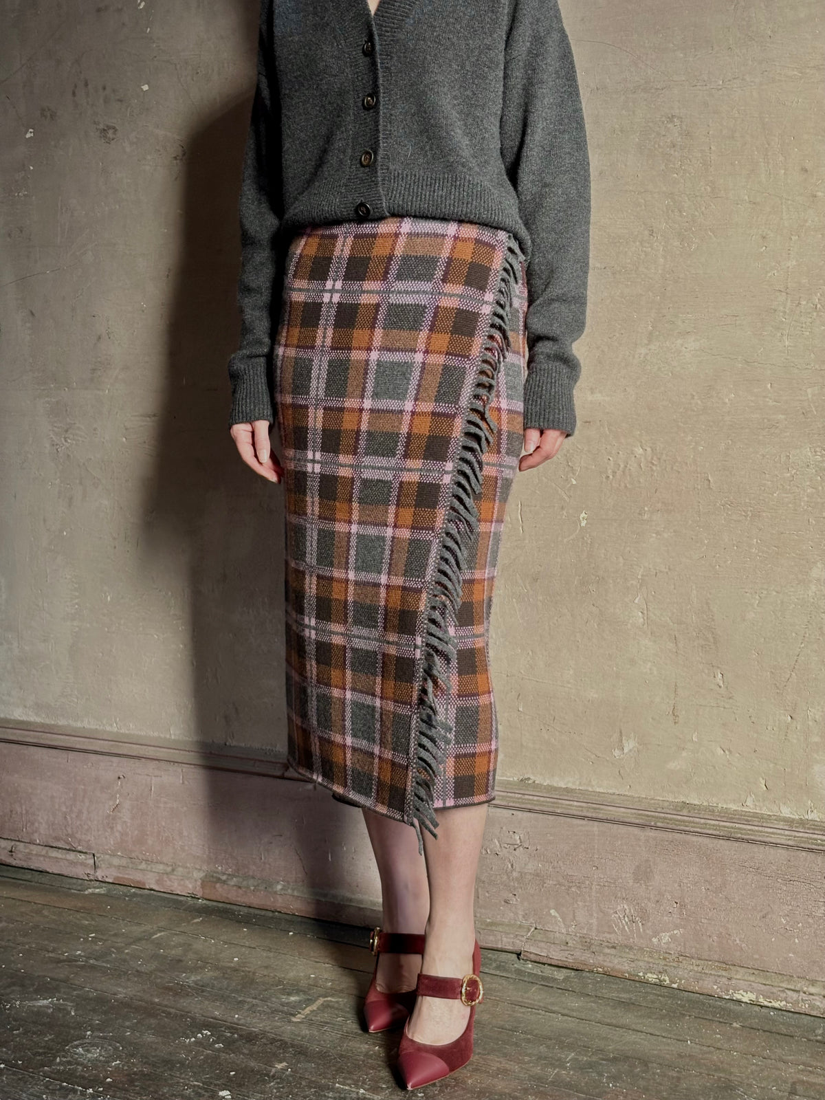 Image of mature woman wearing a gray cardigan with the Guest in Residence Tartan wrap pencil skirt in  brown, orange, and gray plaid with fringe trim 