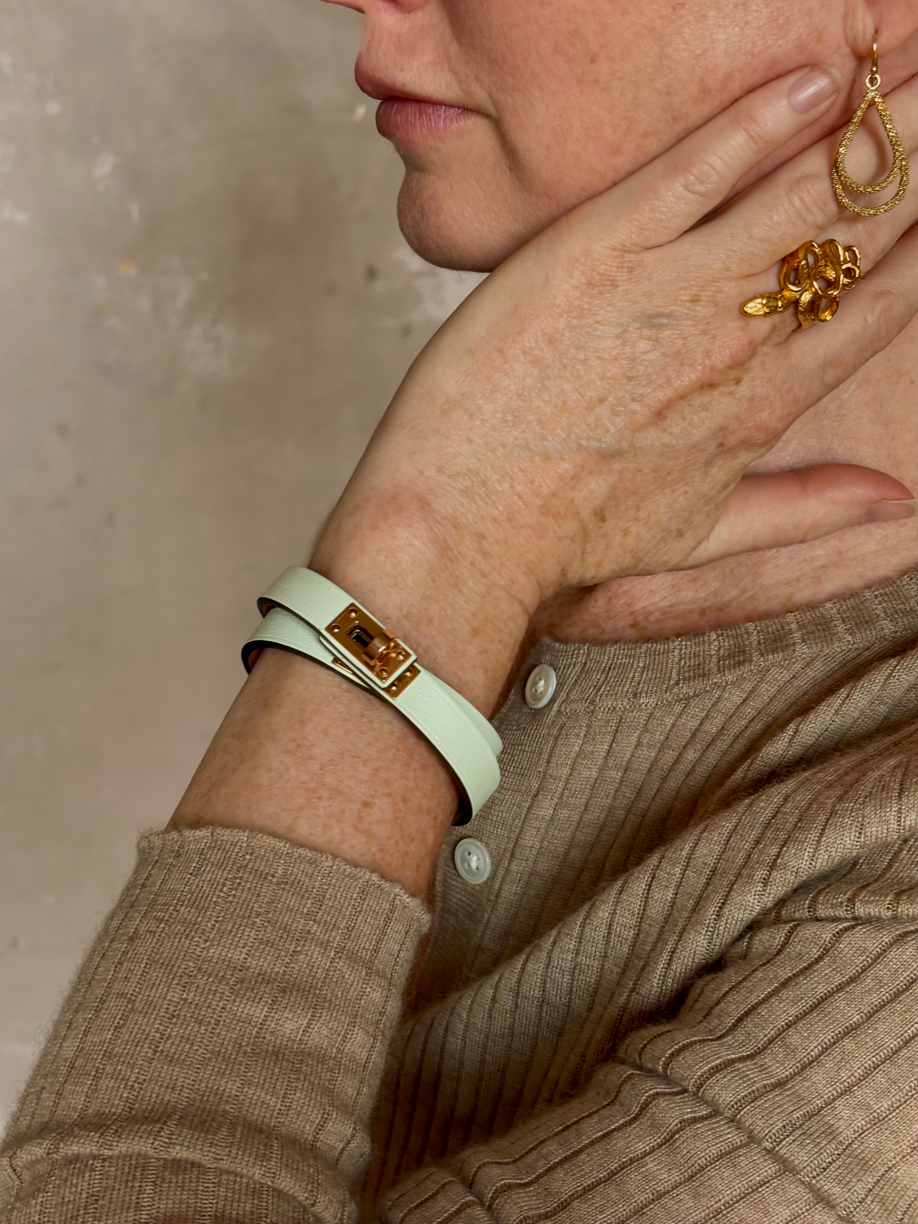 Mature woman wearing Hermès' Mini Kelly Double Tour Wrap Bracelet in Vert d'Eau & Rose Gold pristine, never-worn leather with the iconic Kelly turn-lock. Rare, collectible French luxury