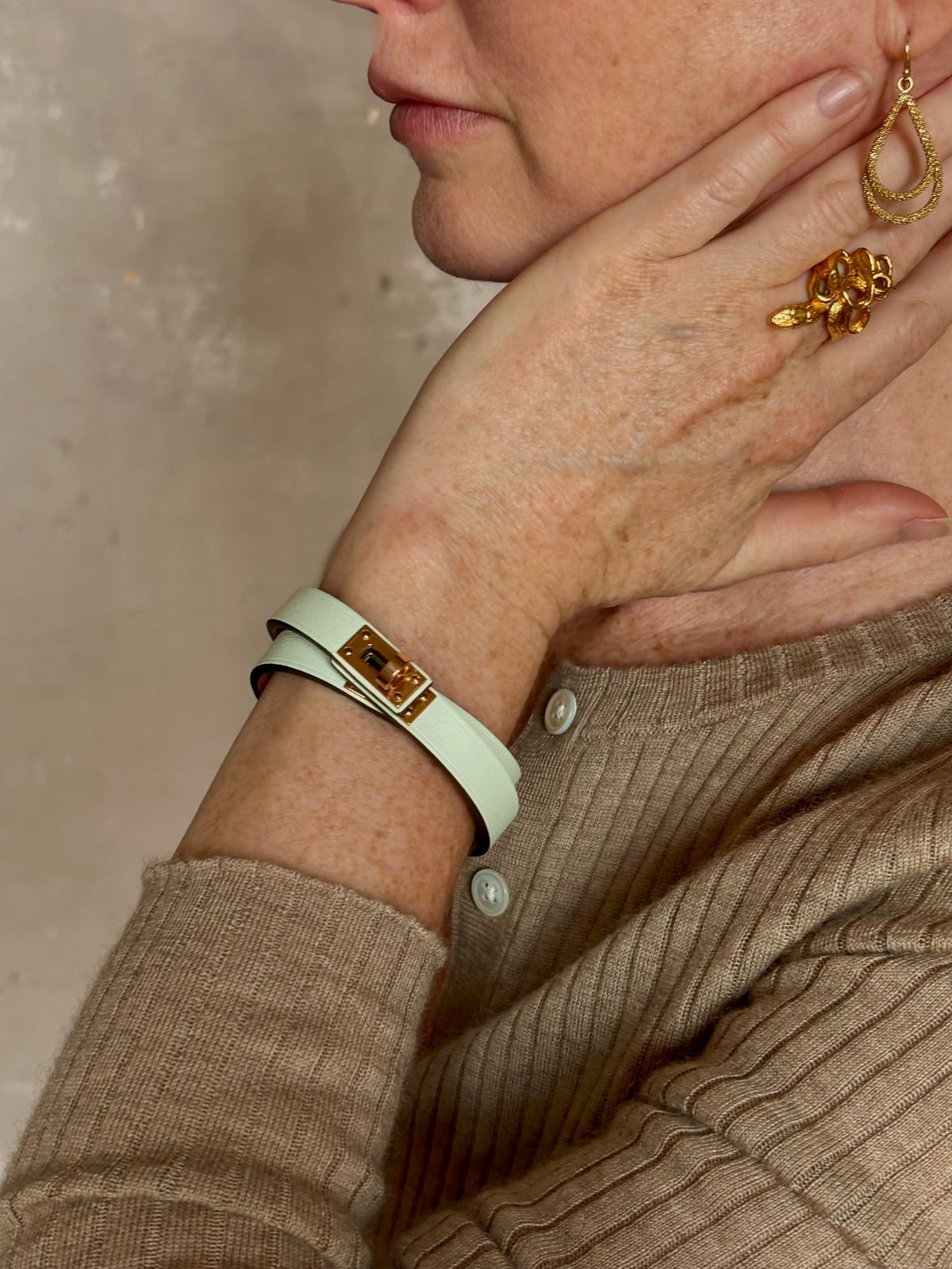 Mature woman wearing Hermès' Mini Kelly Double Tour Wrap Bracelet in Vert d'Eau & Rose Gold pristine, never-worn leather with the iconic Kelly turn-lock. Rare, collectible French luxury