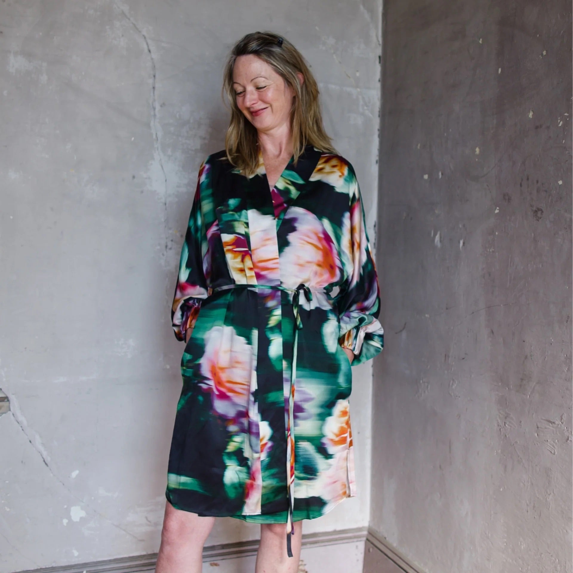 Image featuring a woman wearing the Lunya silk robe designed with waist tie closure, elastic band sleeves and side seam pockets in a bold floral blur print.