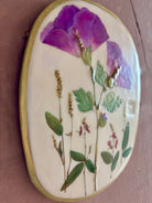 An oval ceramic wall plaque featuring pressed purple rose of sharon hibiscus flowers and green leaves on white clay