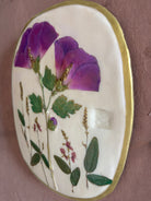 An oval ceramic wall plaque featuring pressed purple rose of sharon hibiscus flowers and green leaves on white clay