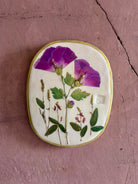 An oval ceramic wall plaque featuring pressed purple rose of sharon hibiscus flowers and green leaves on white clay