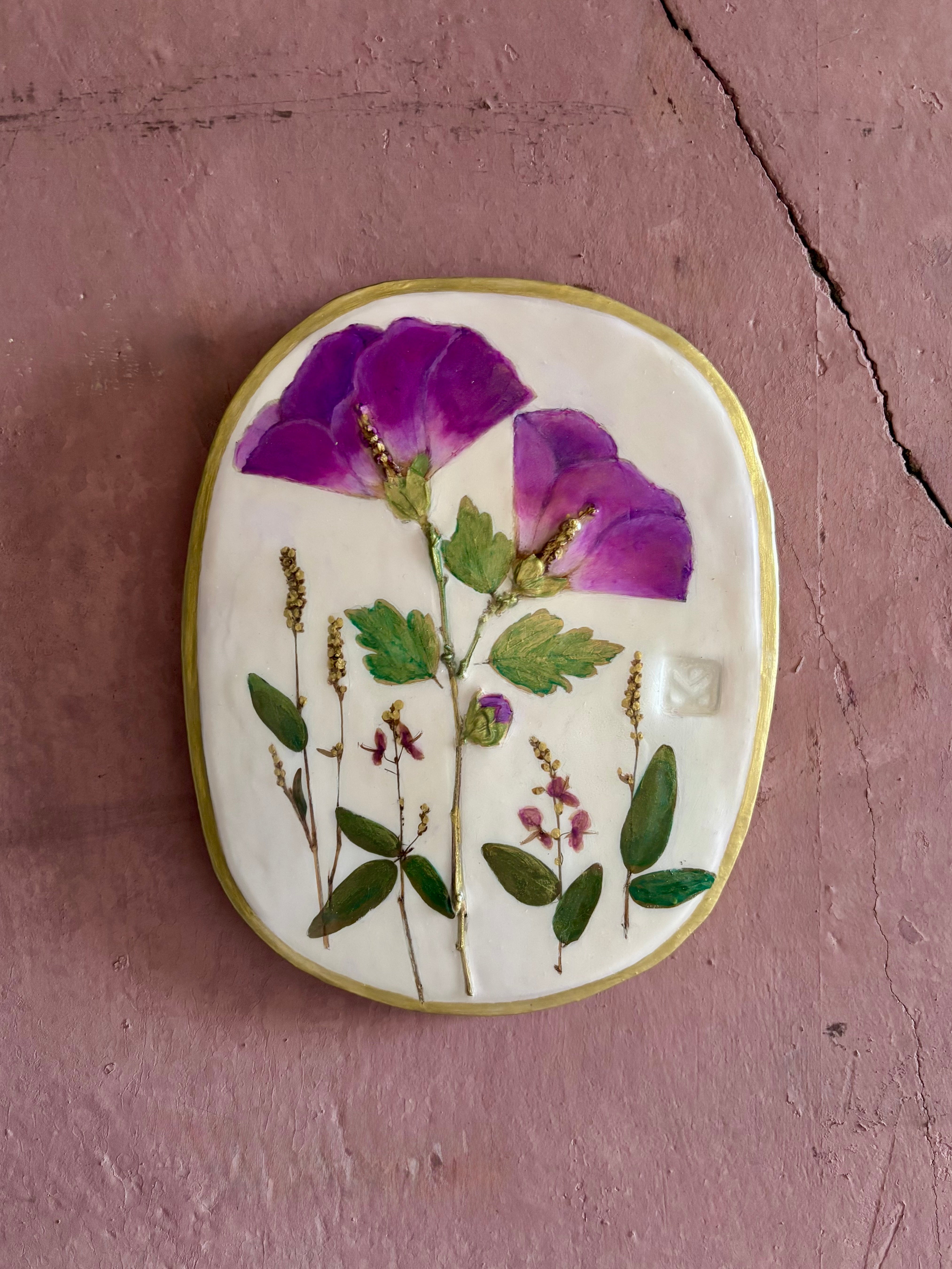 An oval ceramic wall plaque featuring pressed purple rose of sharon hibiscus flowers and green leaves on white clay